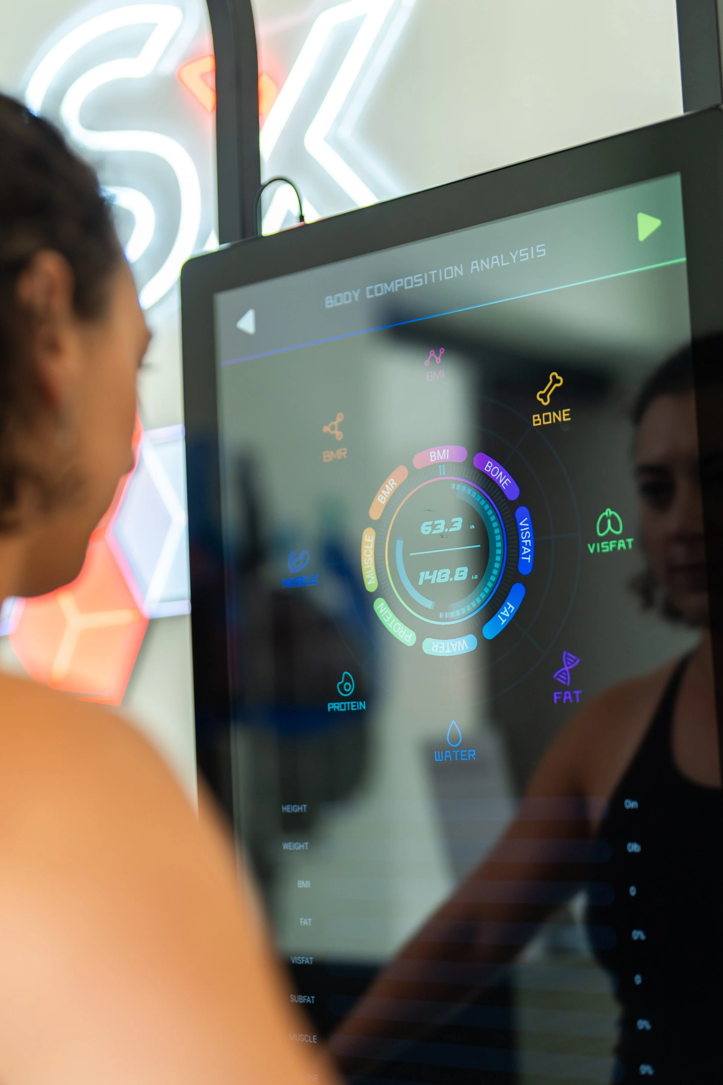 A person viewing a digital screen displaying body composition analysis metrics, including BMI, bone mass, muscle mass, water, and fat percentages, with a neon sign in the background.