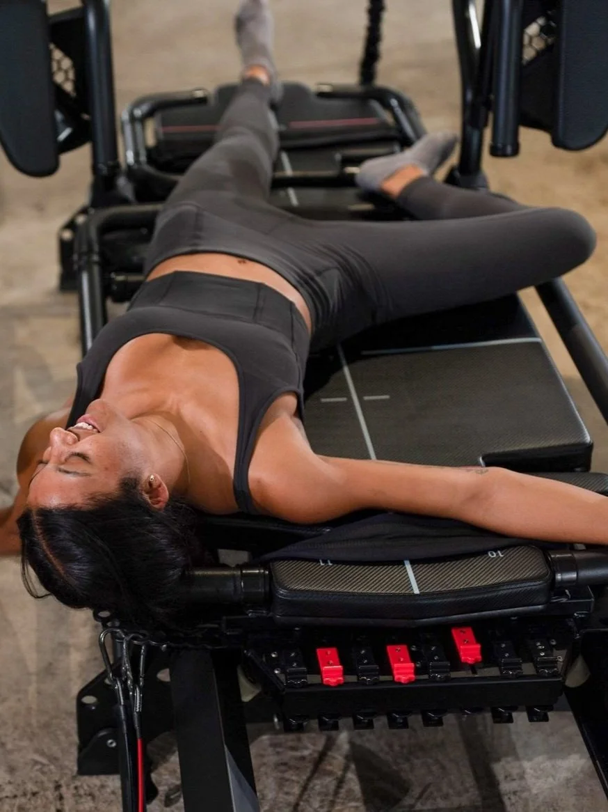 A woman lying on a Pilates reformer machine in a fitness studio, smiling with her eyes closed, wearing black workout clothes.