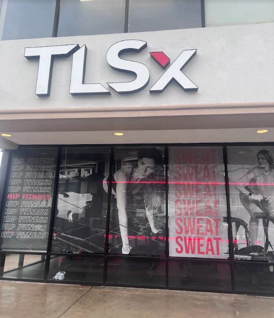 Storefront with large TLSX sign, featuring a gym or fitness center. The window display shows athletic women with text 'SWEAT' repeatedly in pink. The left side has vertical text listing 'HIP FITNESS' multiple times.