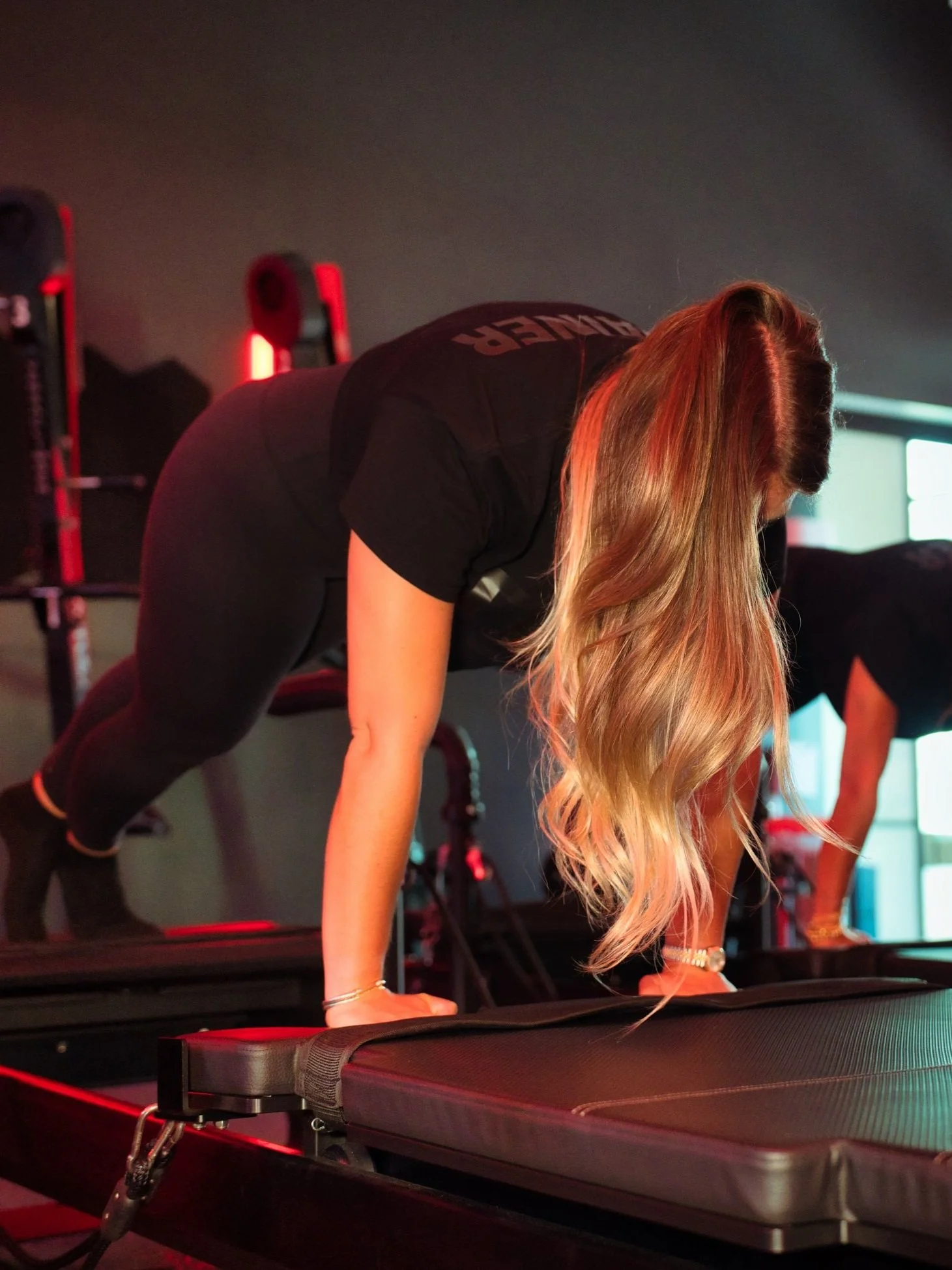 A woman with long, wavy blonde hair is performing a plank exercise on a padded workout platform in a gym, with her hands on the surface and her body parallel to the ground.