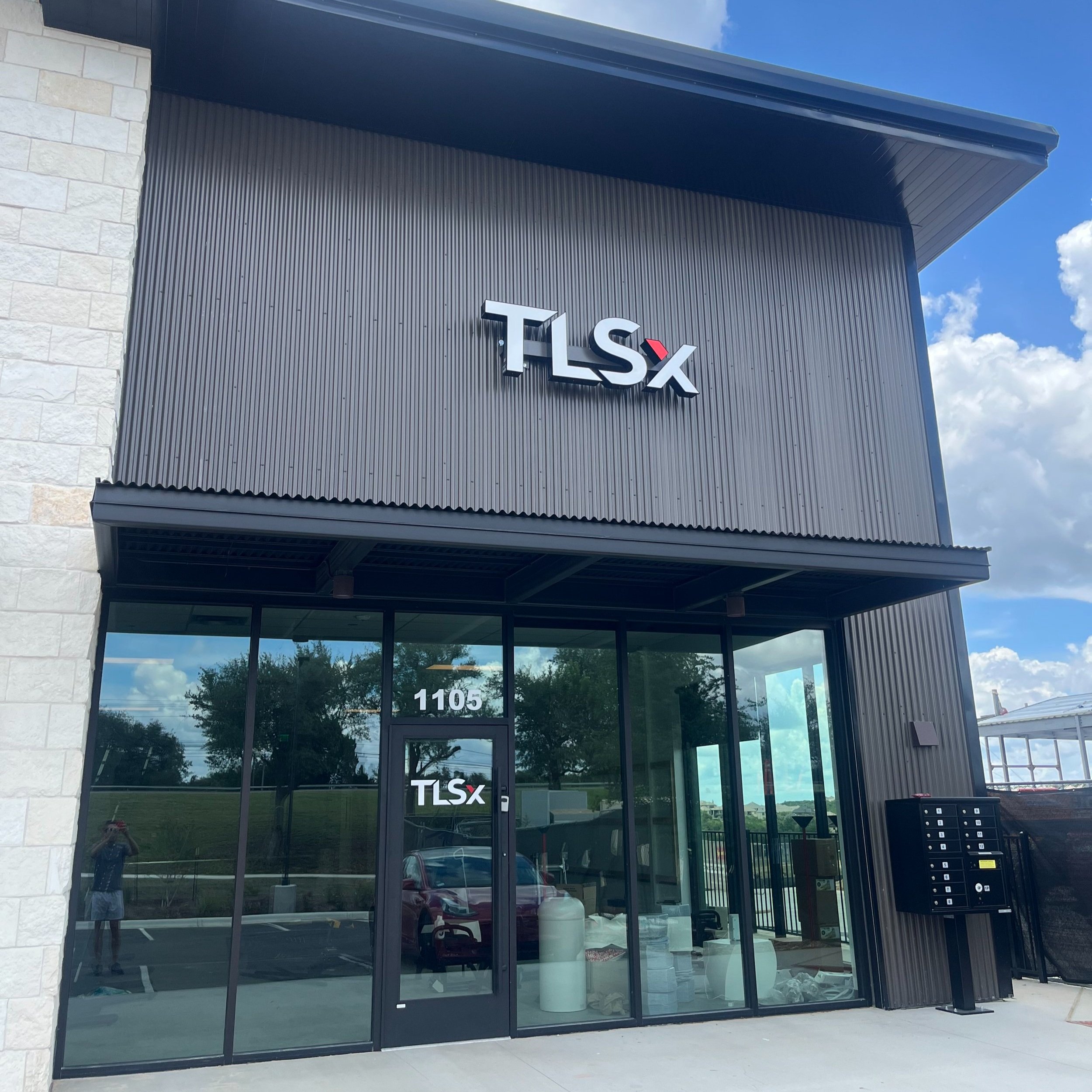 Modern commercial building with black exterior and large glass windows, displaying 'TLSX' signage, with a parking lot and blue sky in the background.