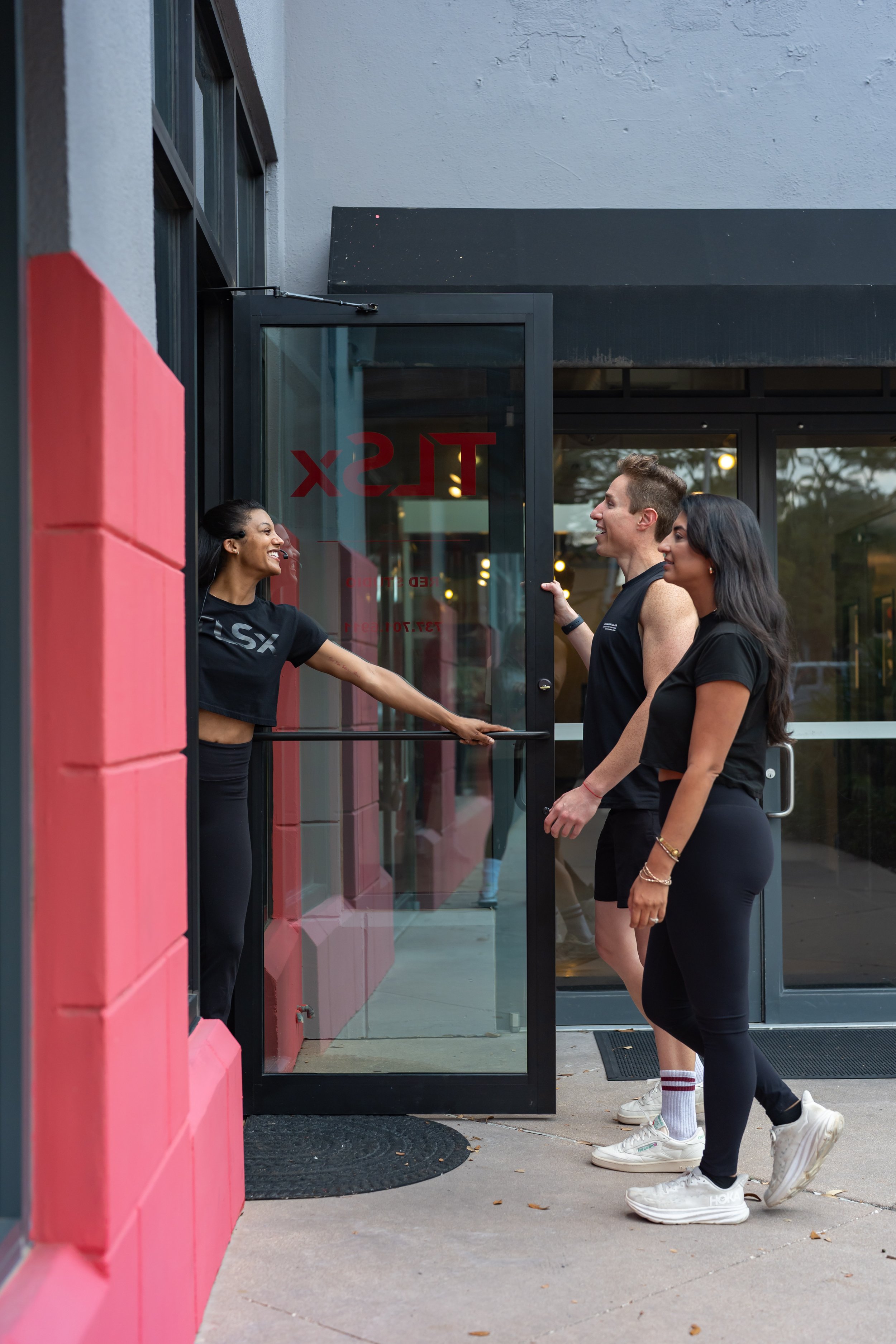A woman standing outside a gym door, holding the door open for a man and woman who are entering, all smiling and wearing workout clothes.