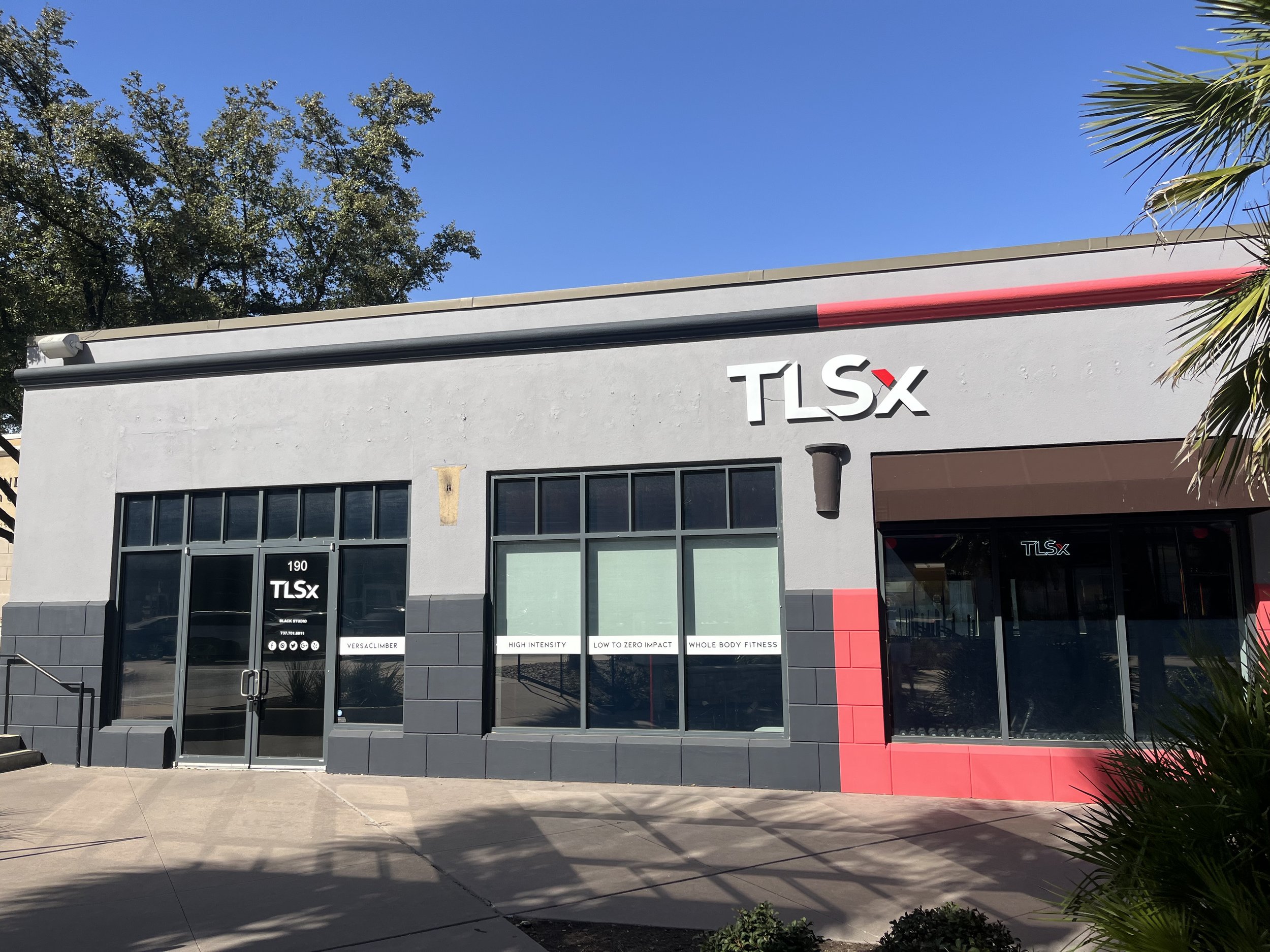 storefront building with TLSx logo, large glass windows, and signs advertising high intensity workouts and whole body fitness, in front of a clear blue sky and trees.