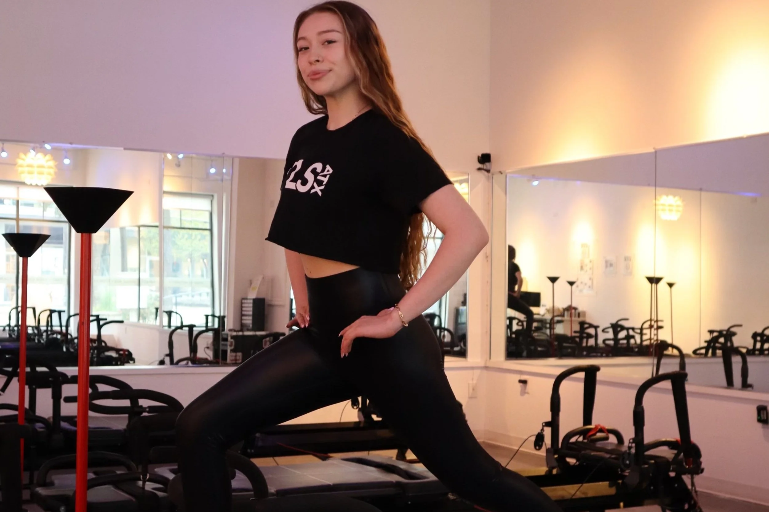 Woman posing in a fitness studio wearing black workout attire, with fitness equipment and mirrors in the background.