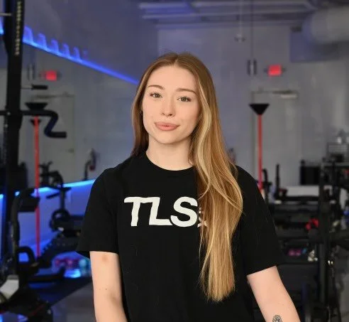 Person with long hair wearing a black TLS T-shirt in a gym setting.
