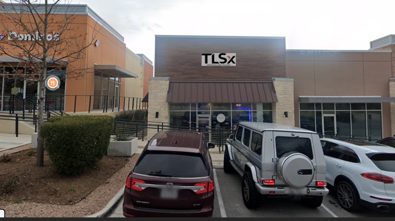 Parking lot in front of a building with the TLSX sign, three parked cars, and a small landscaped area with bushes and trees.