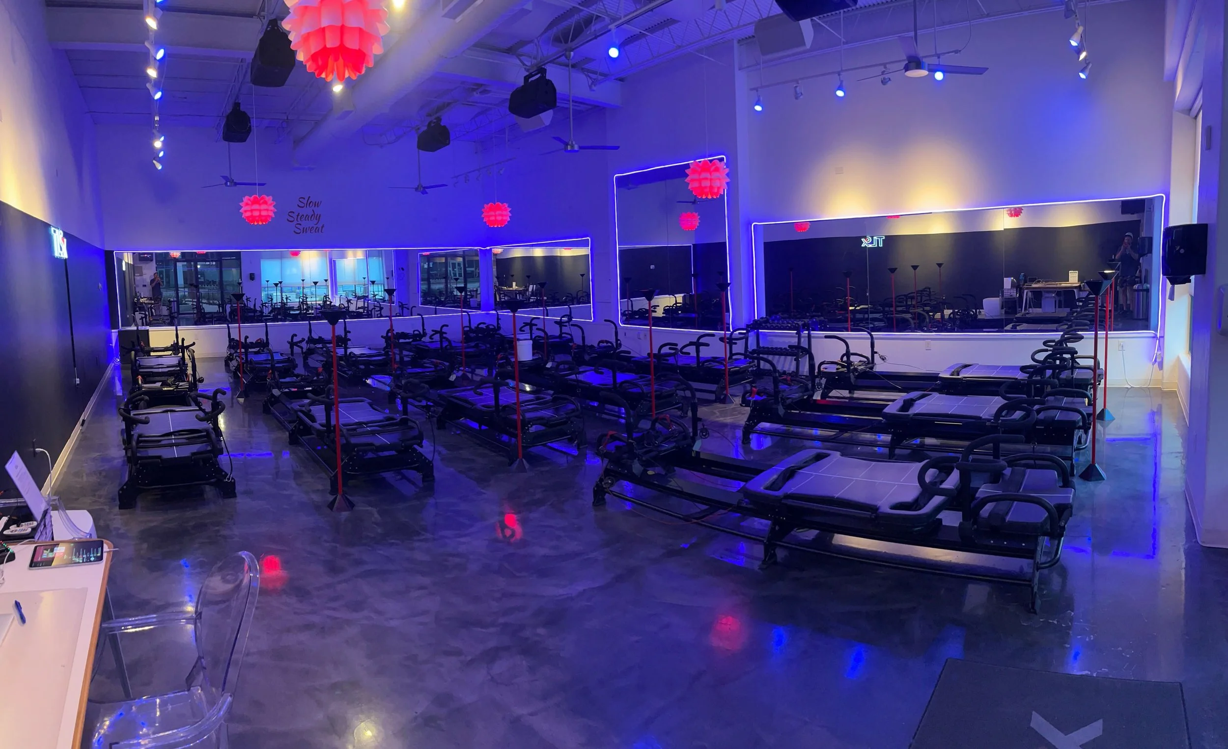Empty pilates studio with rows of reformer and megaformer equipment, purple and blue LED lighting, large mirrors, and red pendant lights hanging from the ceiling.