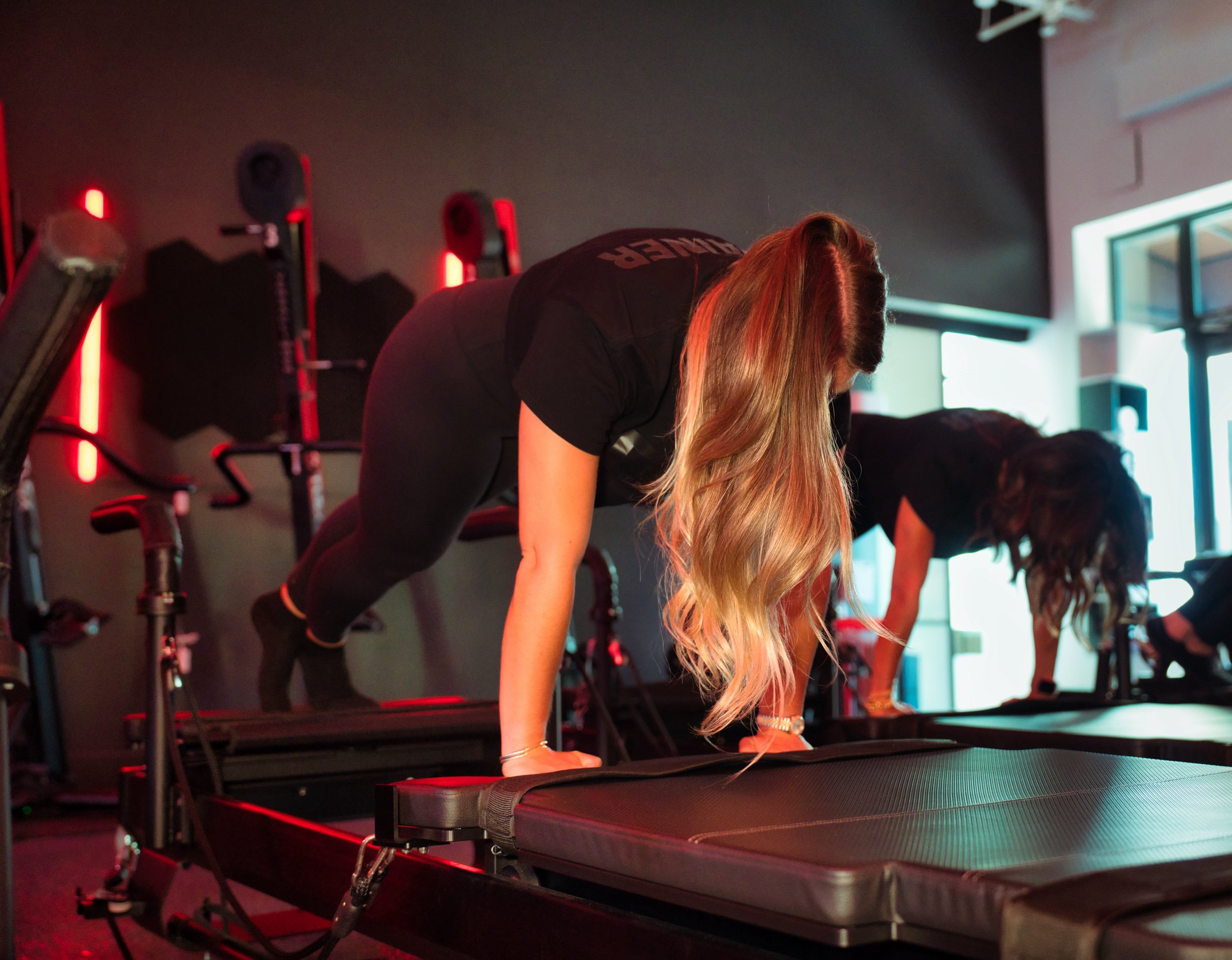 Two women exercising on reformer machines in a gym with black walls and red lighting.