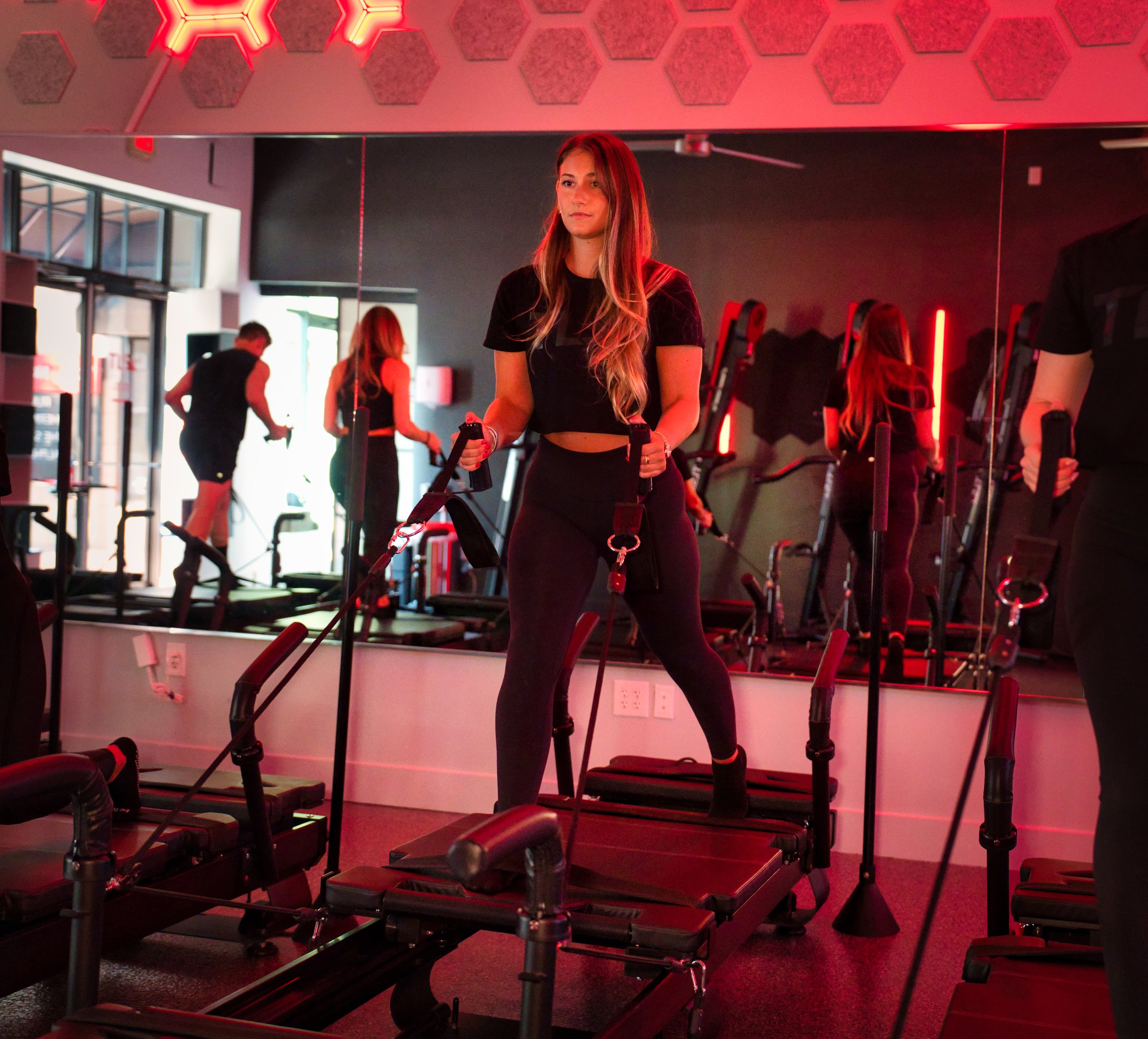 A woman exercising with resistance bands on a Pilates reformer machine in a gym with a red neon light and mirror wall.