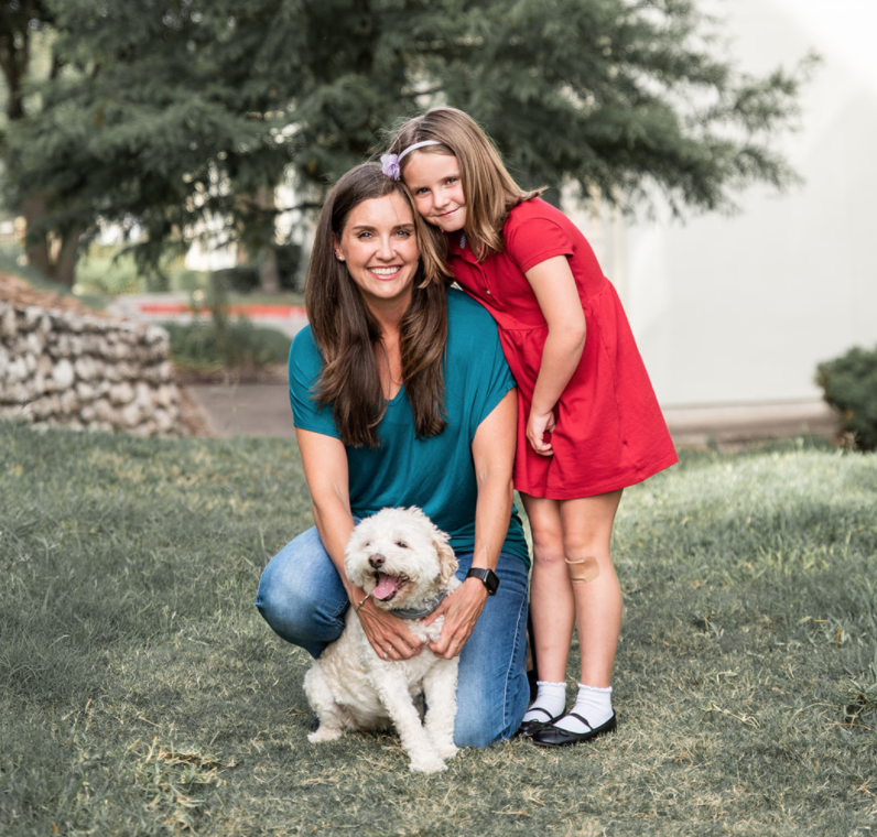 A woman kneeling on grass beside a smiling young girl in a red dress and a white fluffy dog, with trees in the background.