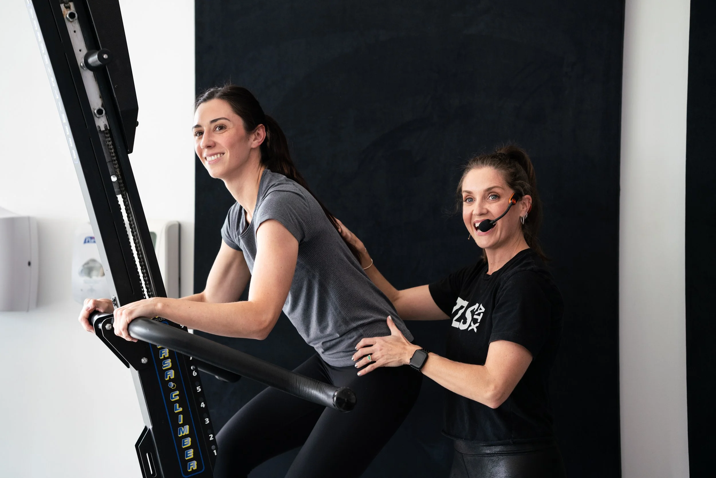 A woman using a VersaClimber exercise machine while an instructor with a headset microphone guides her. The setting appears to be a fitness studio.