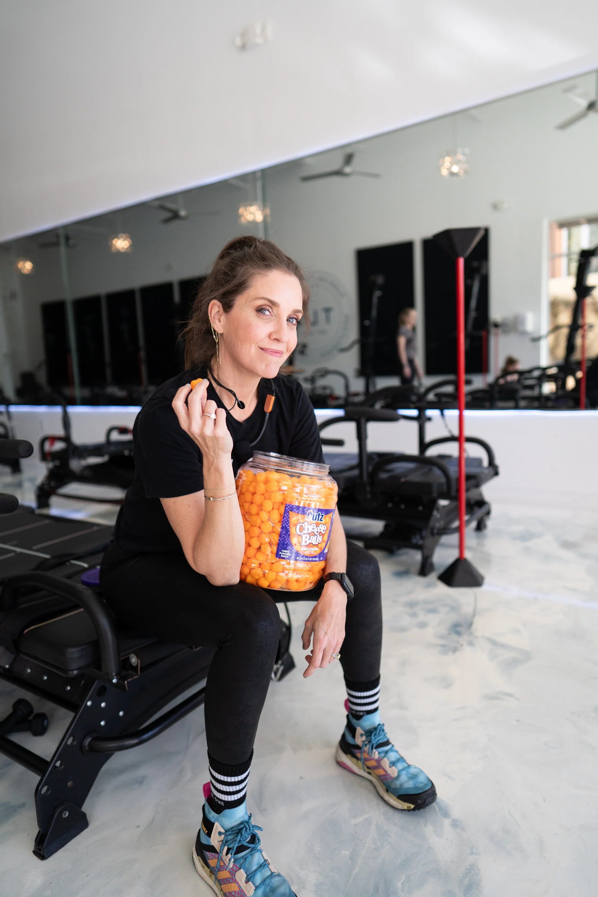 Woman sitting with a jar of cheese balls in a gym setting, wearing sportswear.