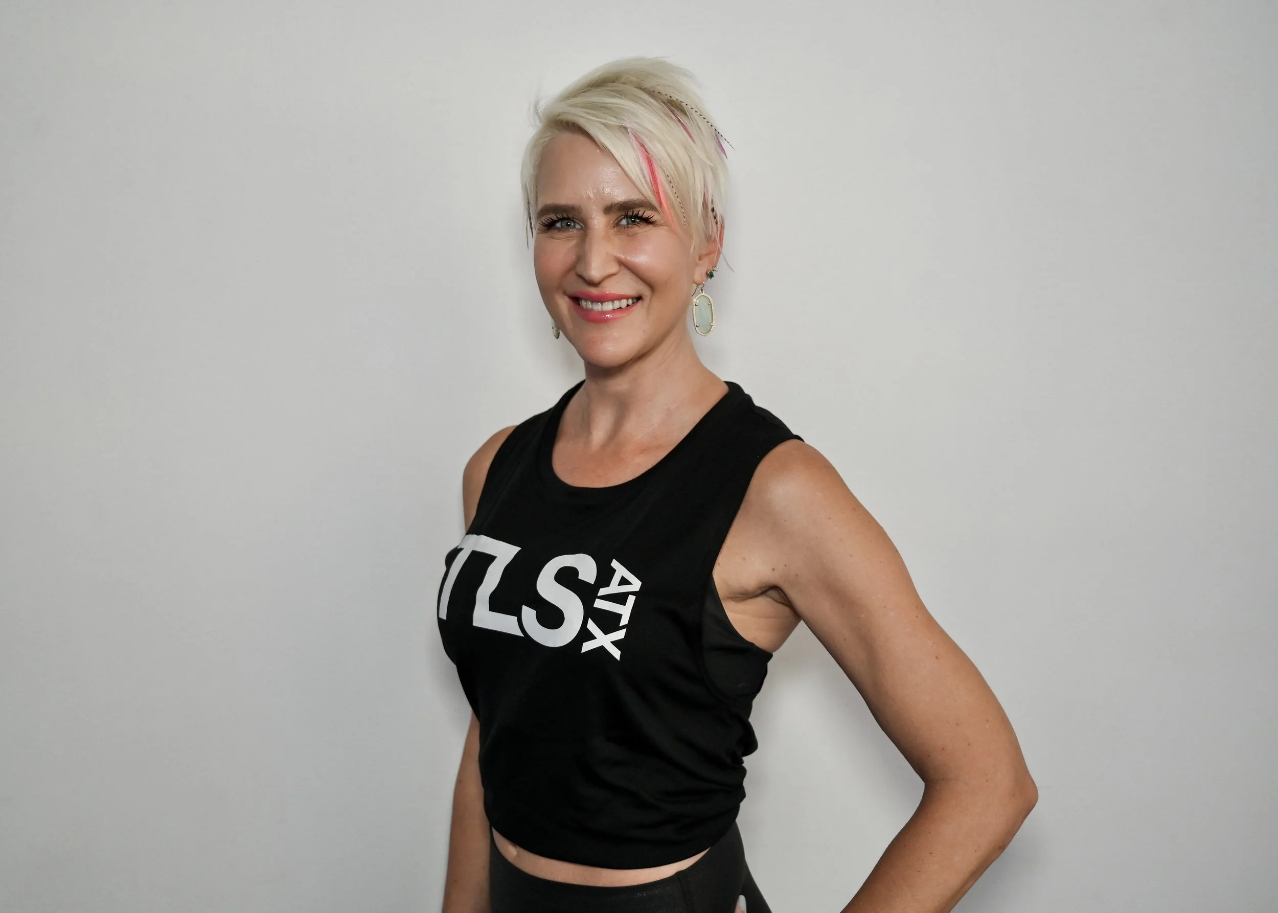 Person with short blonde hair wearing a sleeveless black top with white text "TLS ATX" against a gray background.