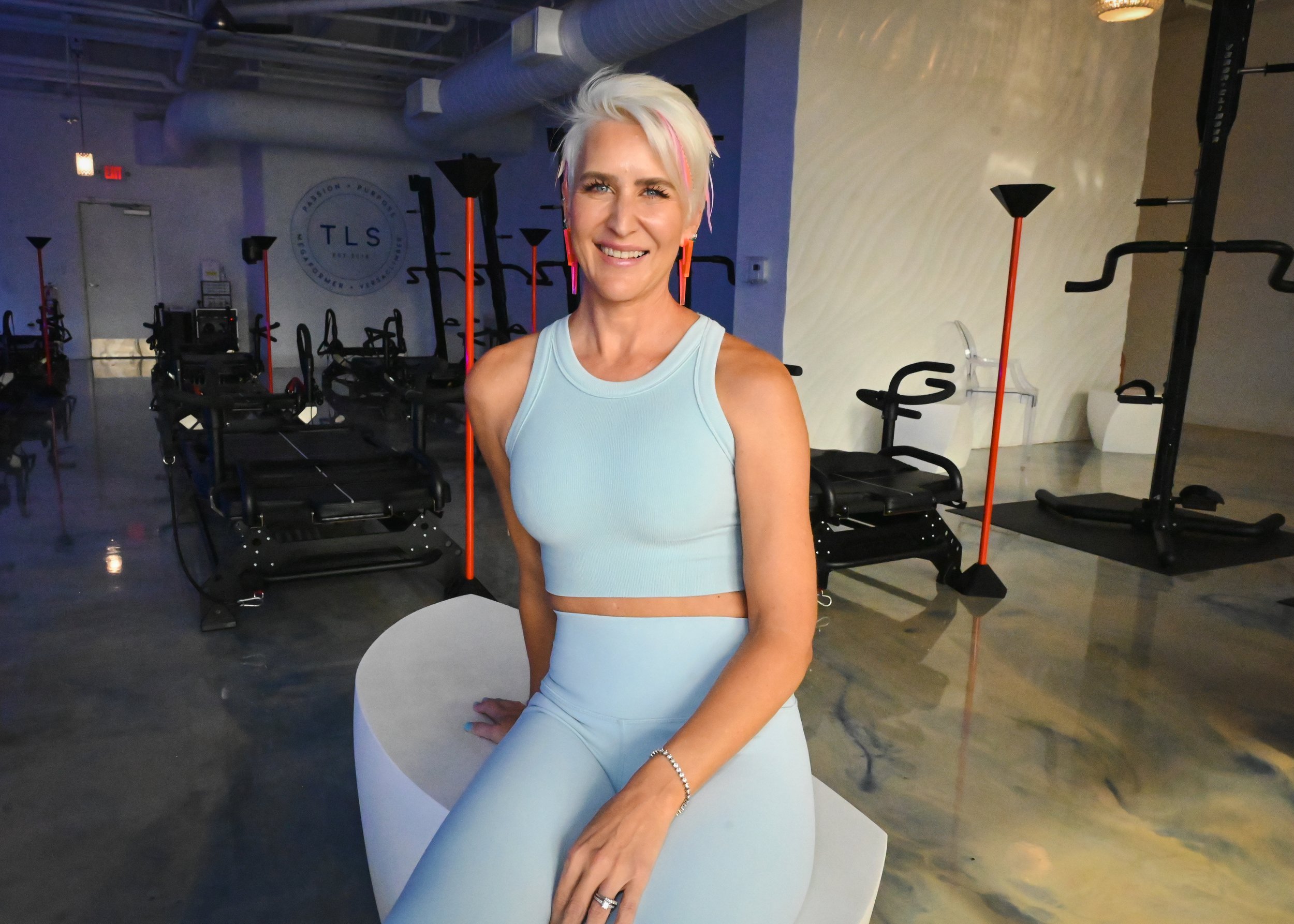 Smiling woman with short blond hair wearing a light blue workout outfit, sitting in a fitness studio with exercise machines in the background.