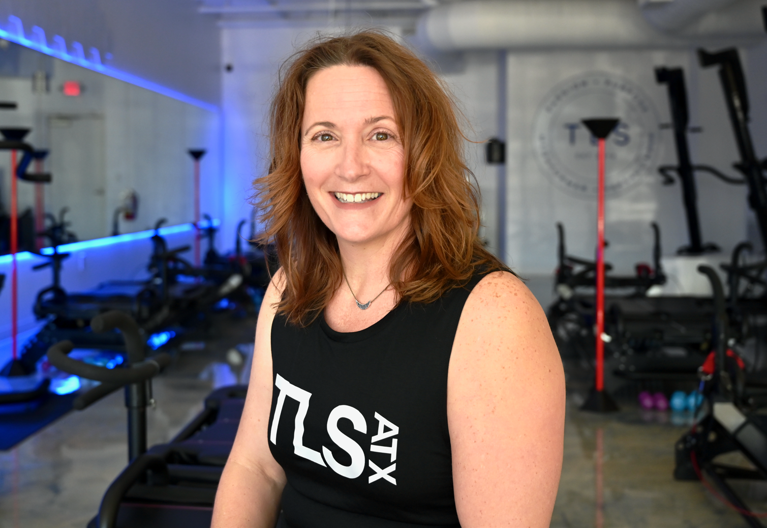 Woman smiling inside a fitness studio with gym equipment and a "TLS ATX" tank top.