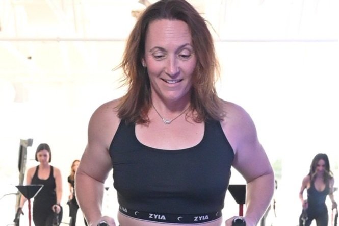 A woman in a black sports bra exercising on gym equipment with others in the background.