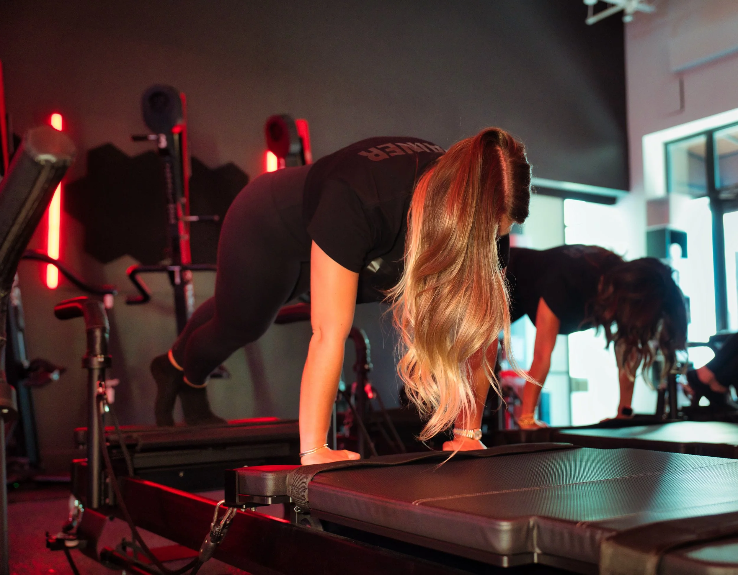Two women exercising on reformer machines in a gym with black walls and red lighting.