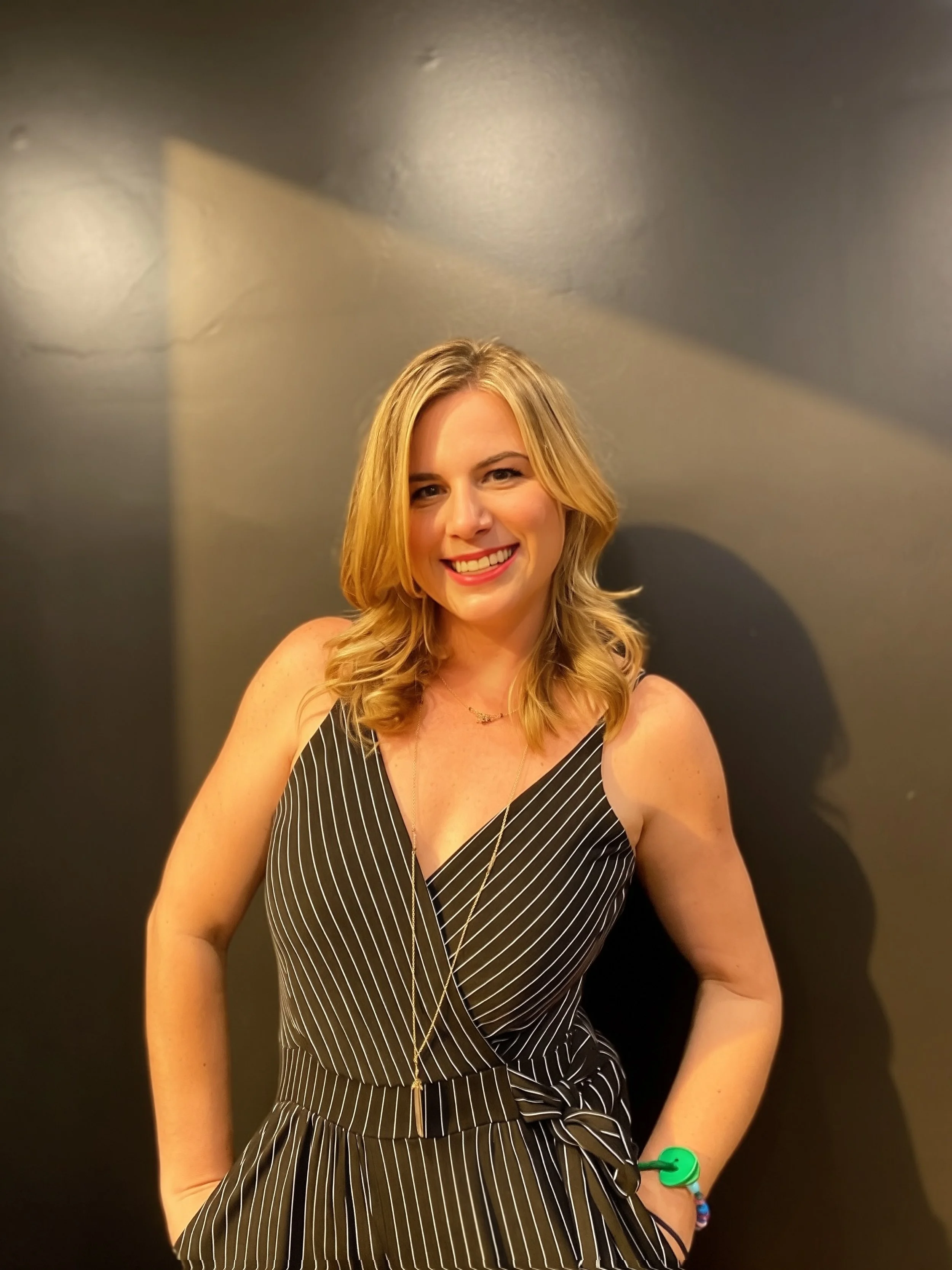 A woman with blonde hair in loose waves, wearing a black and white striped sleeveless dress, smiling and standing against a plain dark gray wall.