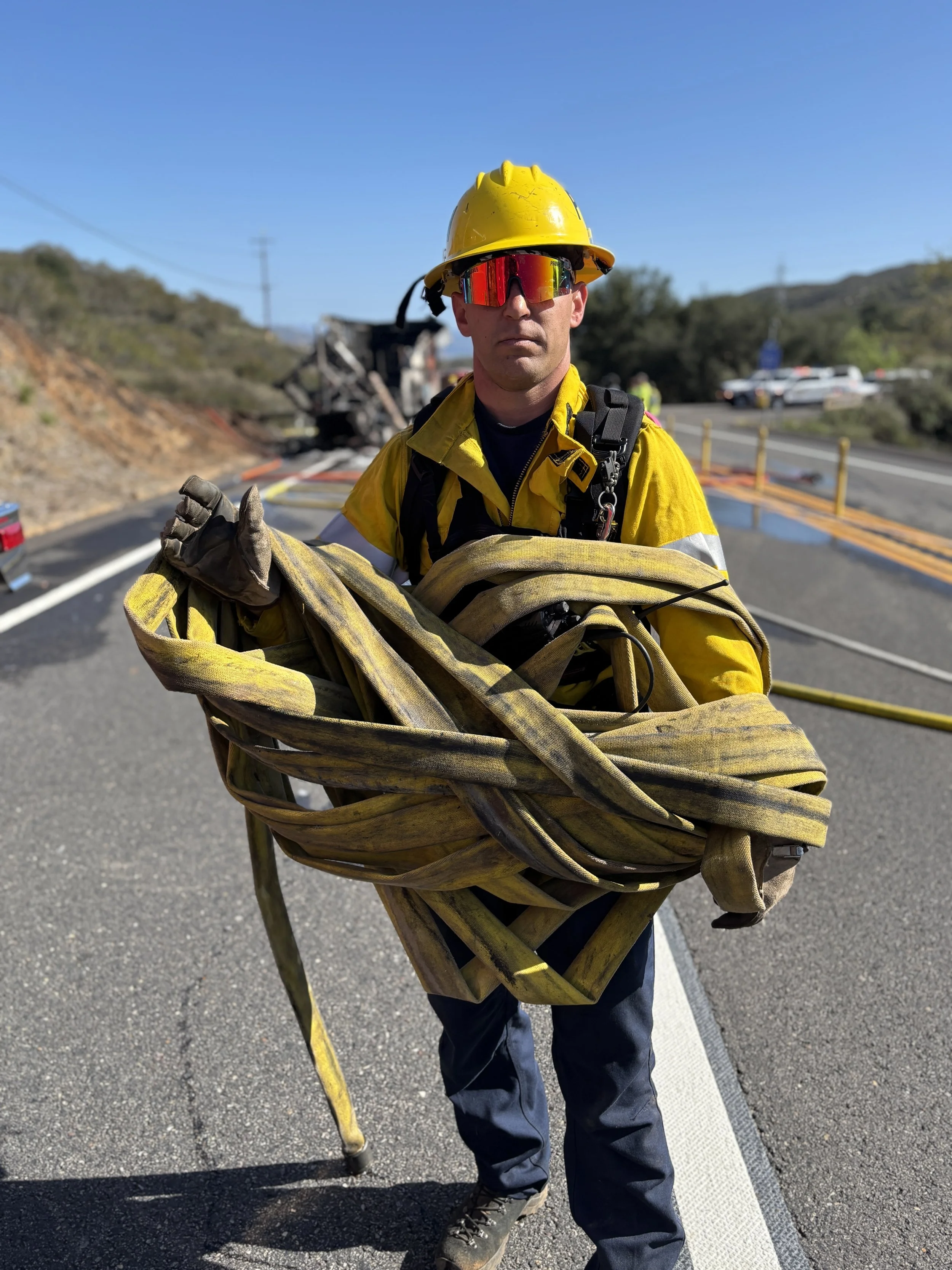 Firefighter in yellow uniform and helmet holding a rolled-up fire hose on the side of a highway after a car accident.
