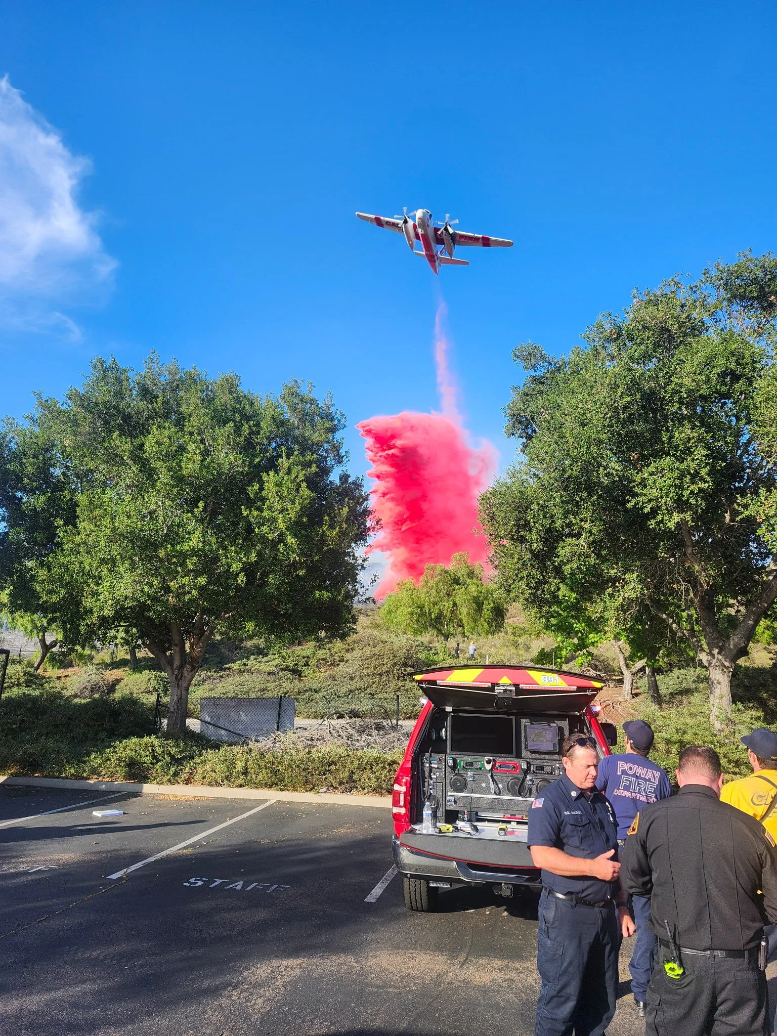 Firefighters and emergency personnel observe a firefighting aircraft releasing red fire retardant over a wildfire, with a group of trees and a blue sky in the background.