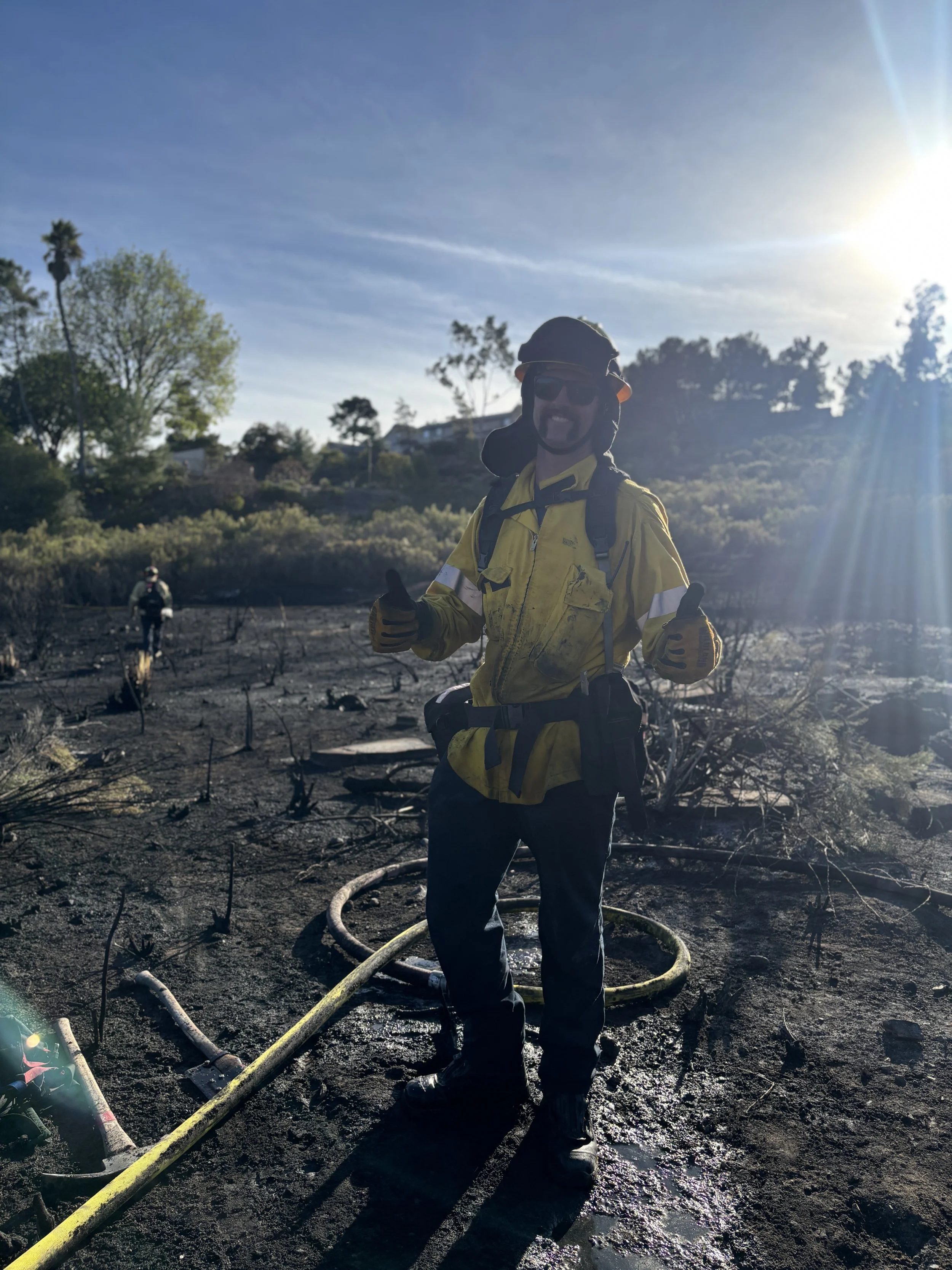 A man in firefighter gear, smiling and giving a thumbs-up, standing on a burnt, charred landscape with another person in the background, under a cloudy sky with the sun shining brightly.