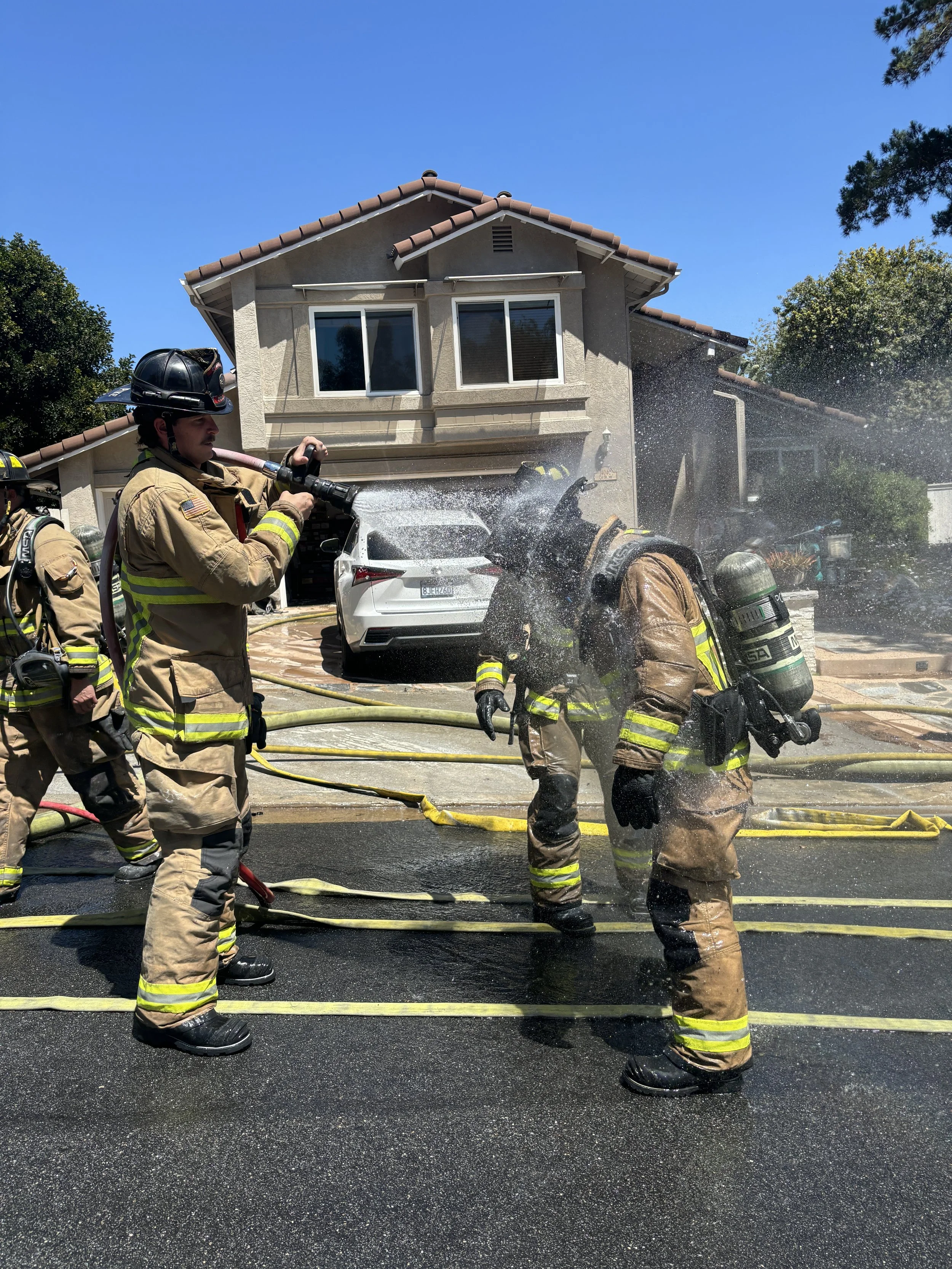 Firefighters actively spraying water on a house fire on a suburban street during daytime, with a white car parked in front of the house.