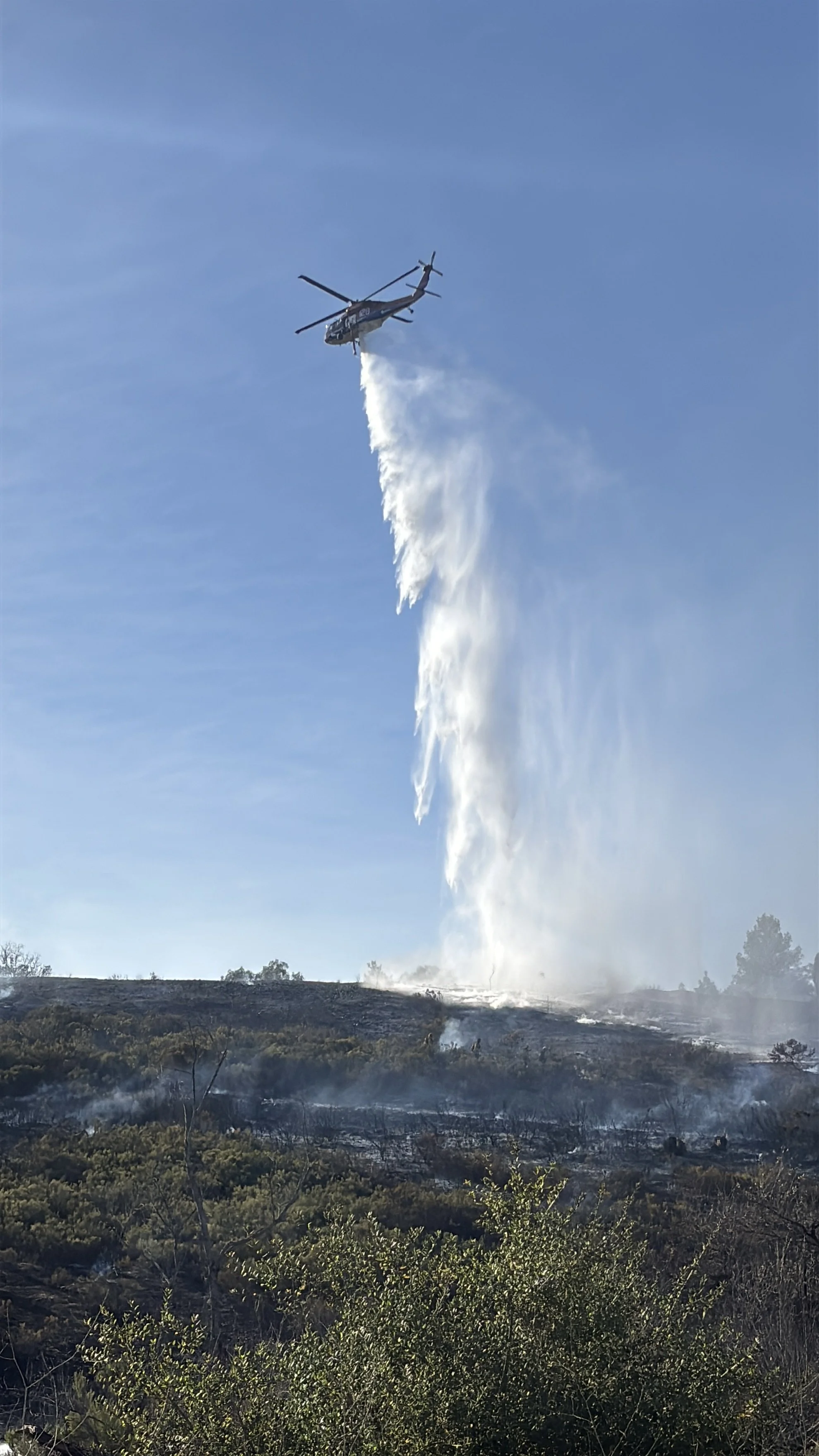 Helicopter dropping water on a wildfire in a forested area.