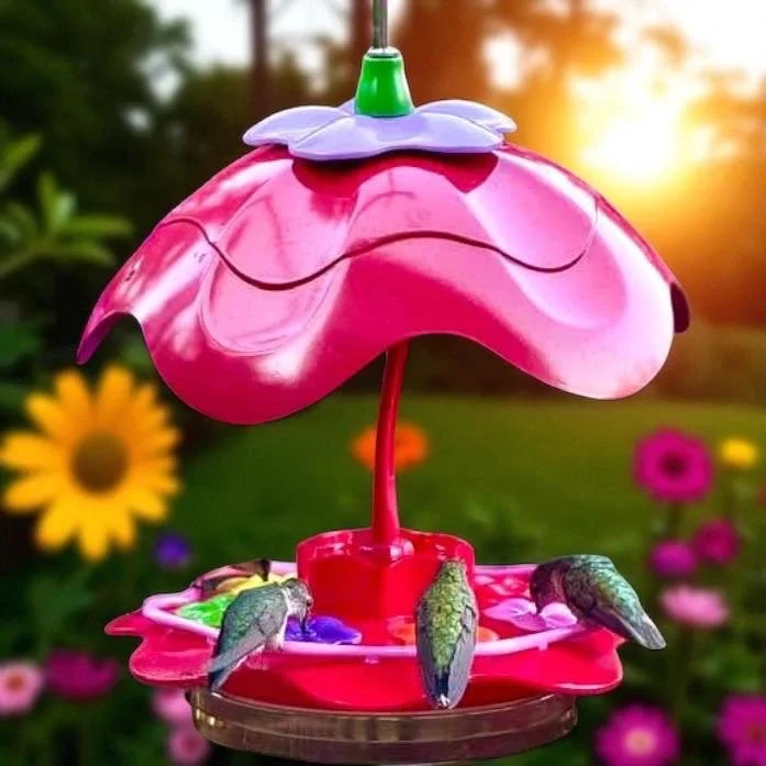 Colorful hummingbird feeder with pink flowers, set against a garden background with blooming flowers and a sunset.