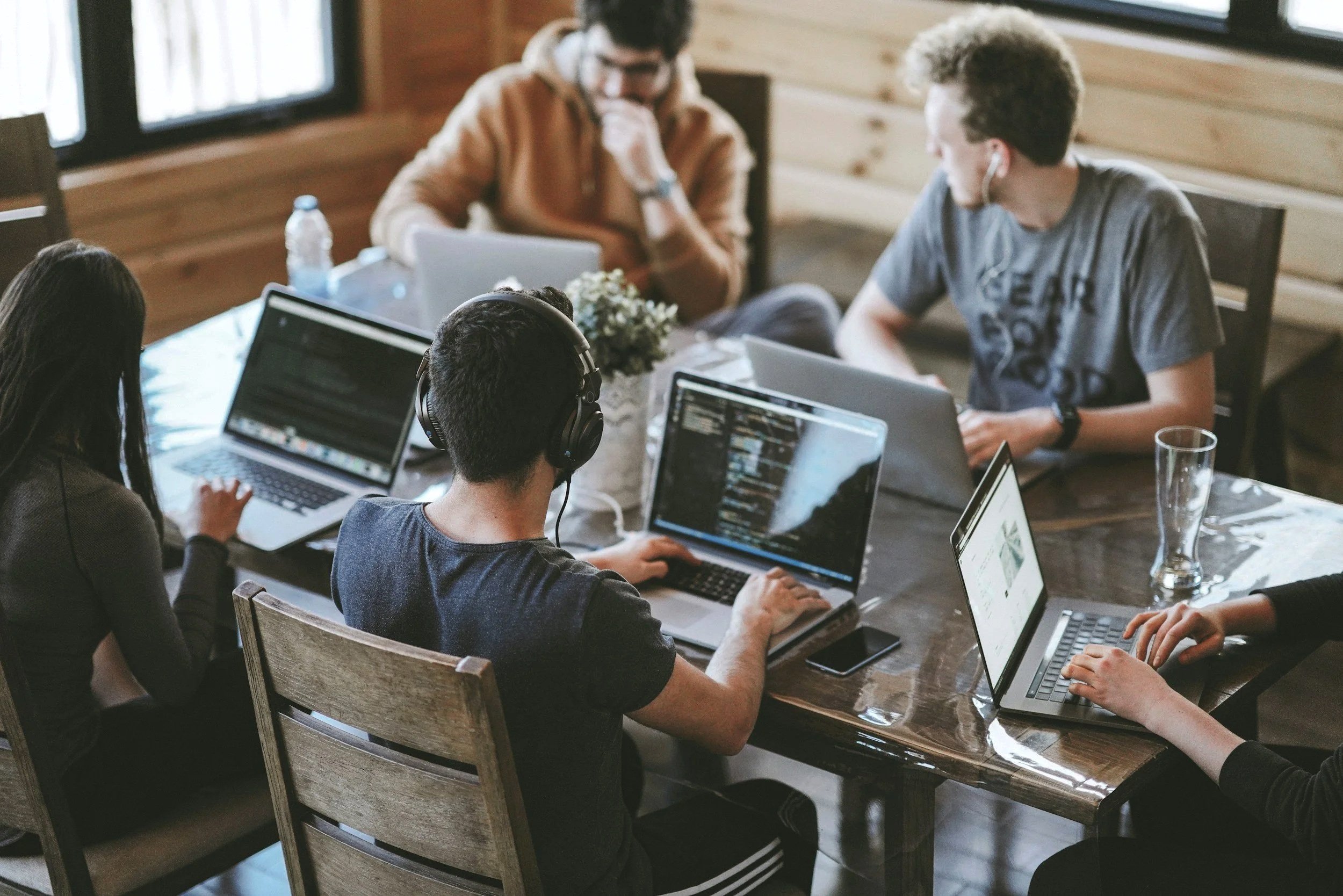 Grupo de jóvenes trabajando en una reunión en una mesa de madera, usando computadoras portátiles y con una decoración de madera en las paredes.