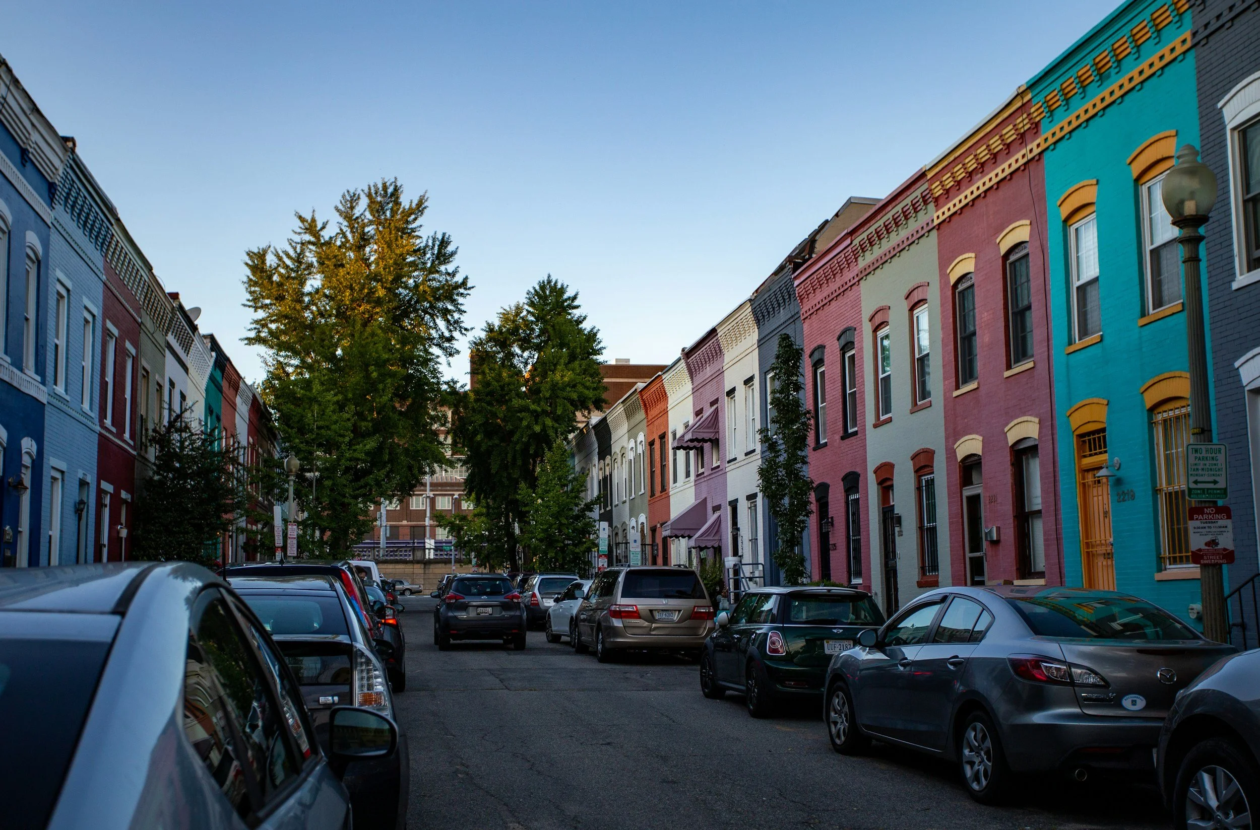 Colorful row of townhouses, in Washington D.C. with parked cars along a street and trees in the background on a clear day.