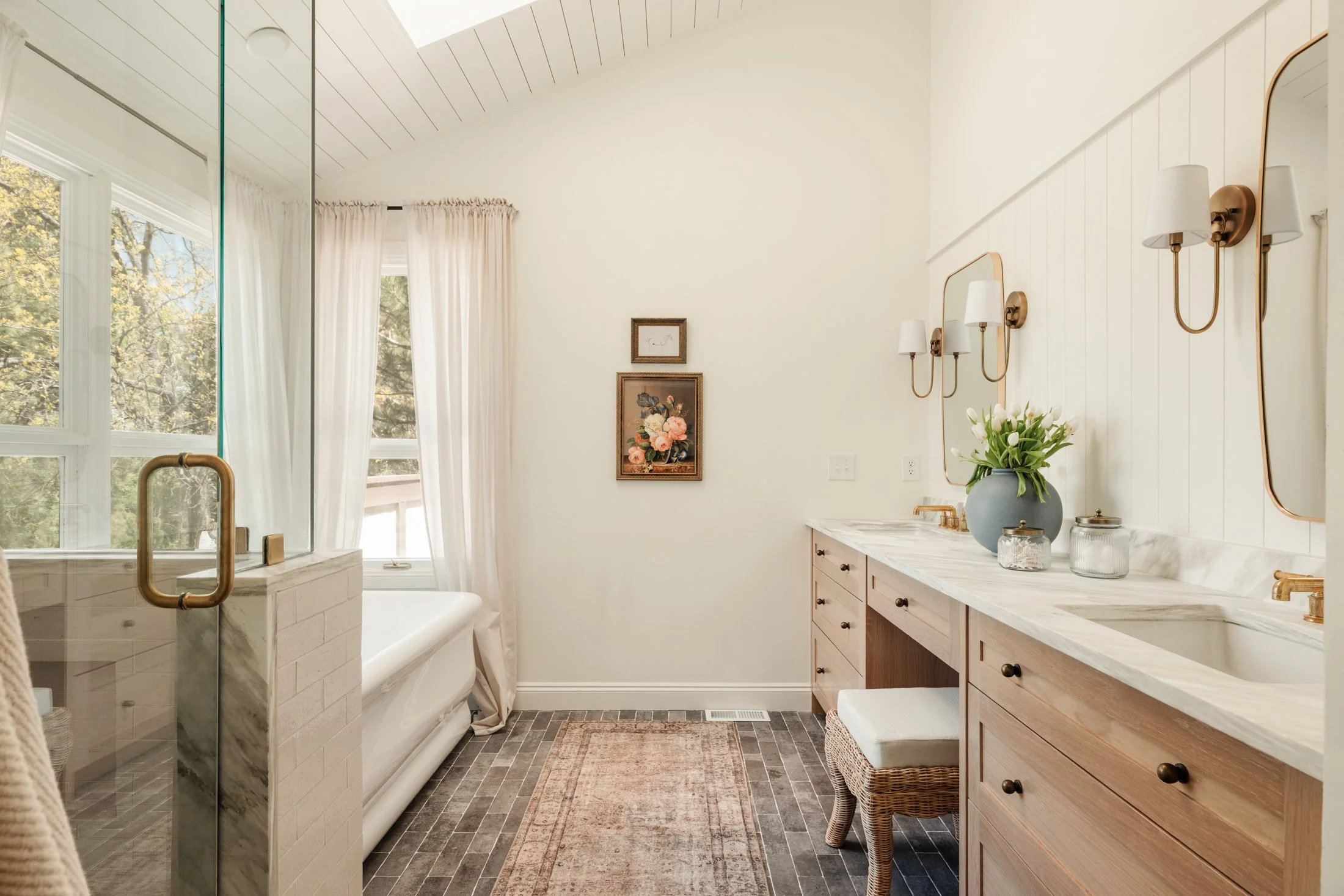 Bathroom with white walls, wooden vanity, marble countertop, wall-mounted light fixtures, a vase of white tulips, artwork, and a window with white curtains showing trees outside.