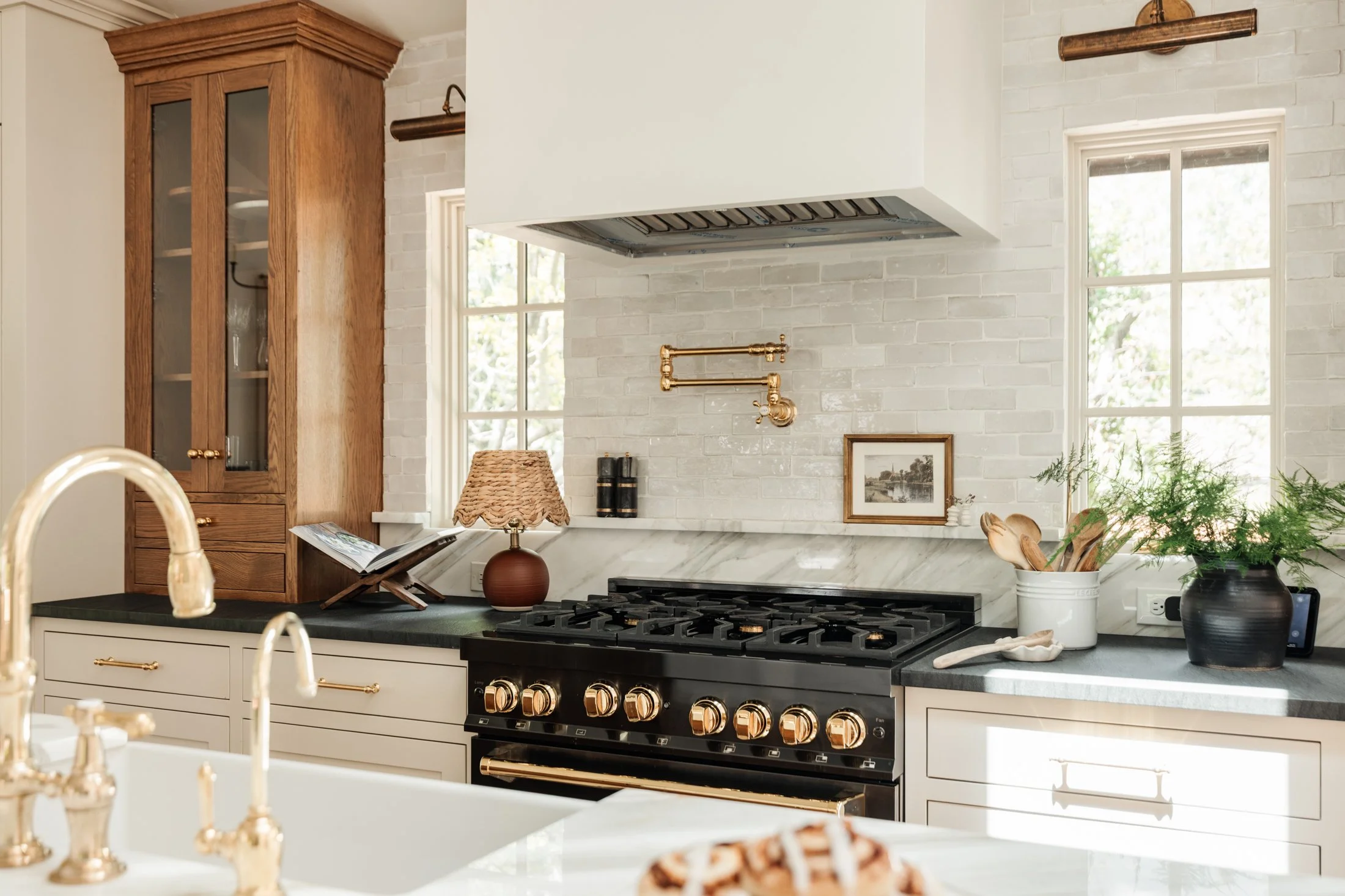 Kitchen with white brick wall, wooden cabinetry, gas stove, and window with outdoor view. Elements include a copper pipe, framed picture, potted plant, and a woven lampshade.