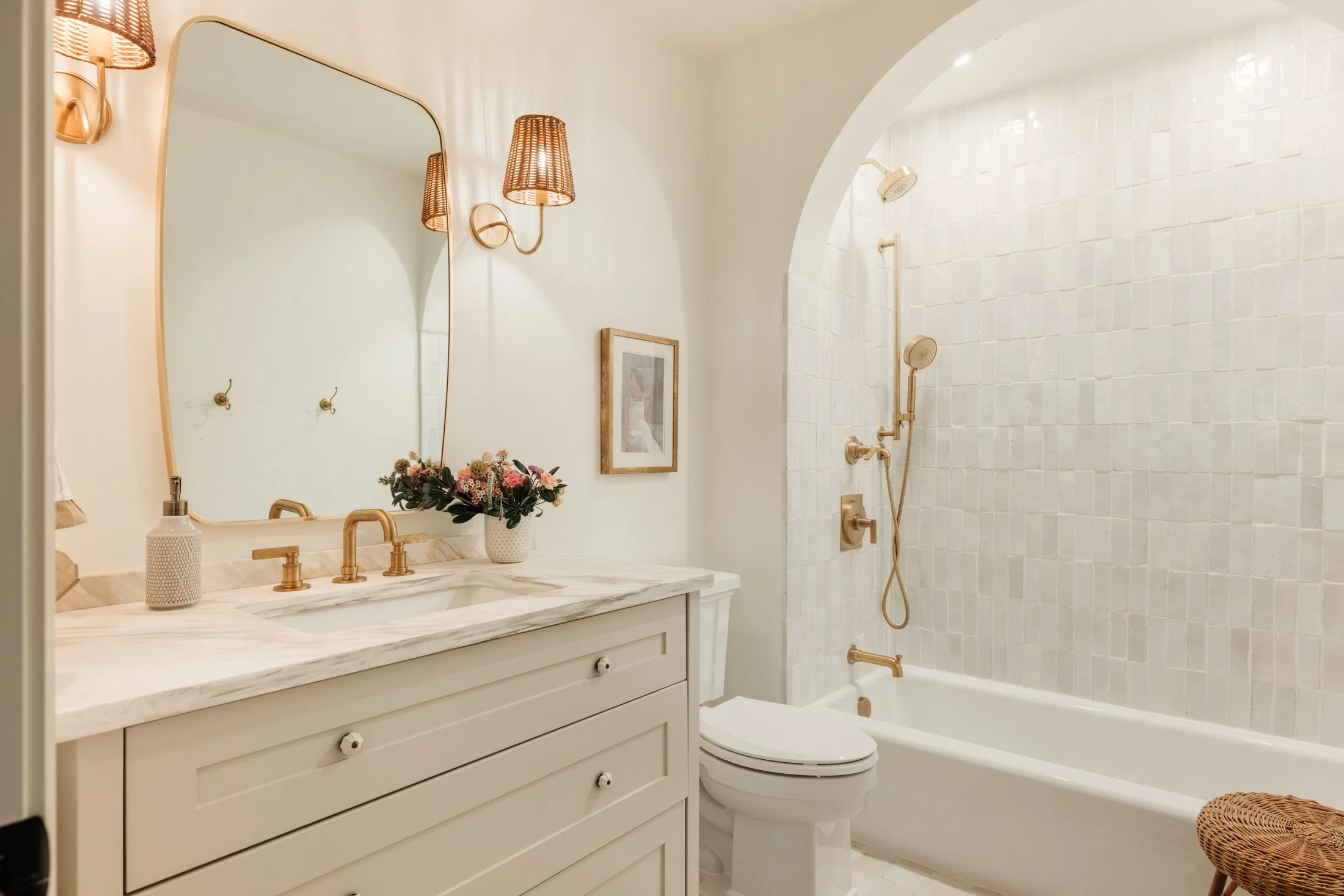 A modern bathroom with a white vanity, a marble countertop, brass fixtures, and a bouquet of flowers. The bathroom has a large mirror, a framed picture, and wicker wall sconces. There is a bathtub with a showerhead and brass fixtures visible in the background.