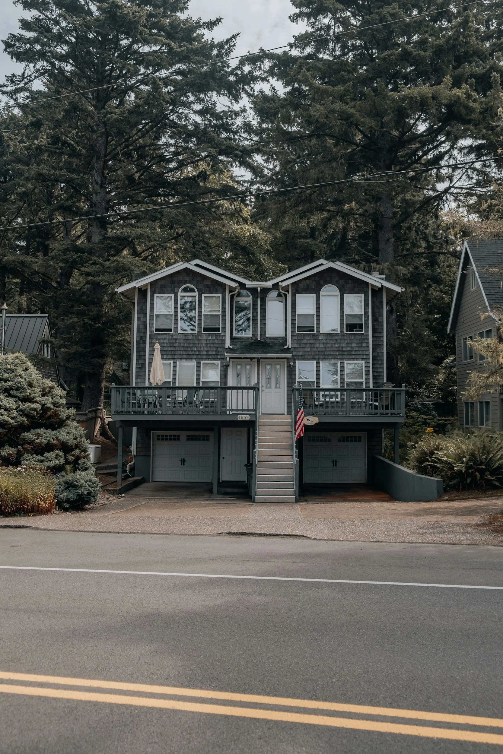 A multi-story house with a front deck, stairs, and garage, set against a background of tall evergreen trees. The house has grey shingles, white window frames, and a small American flag near the stairs.