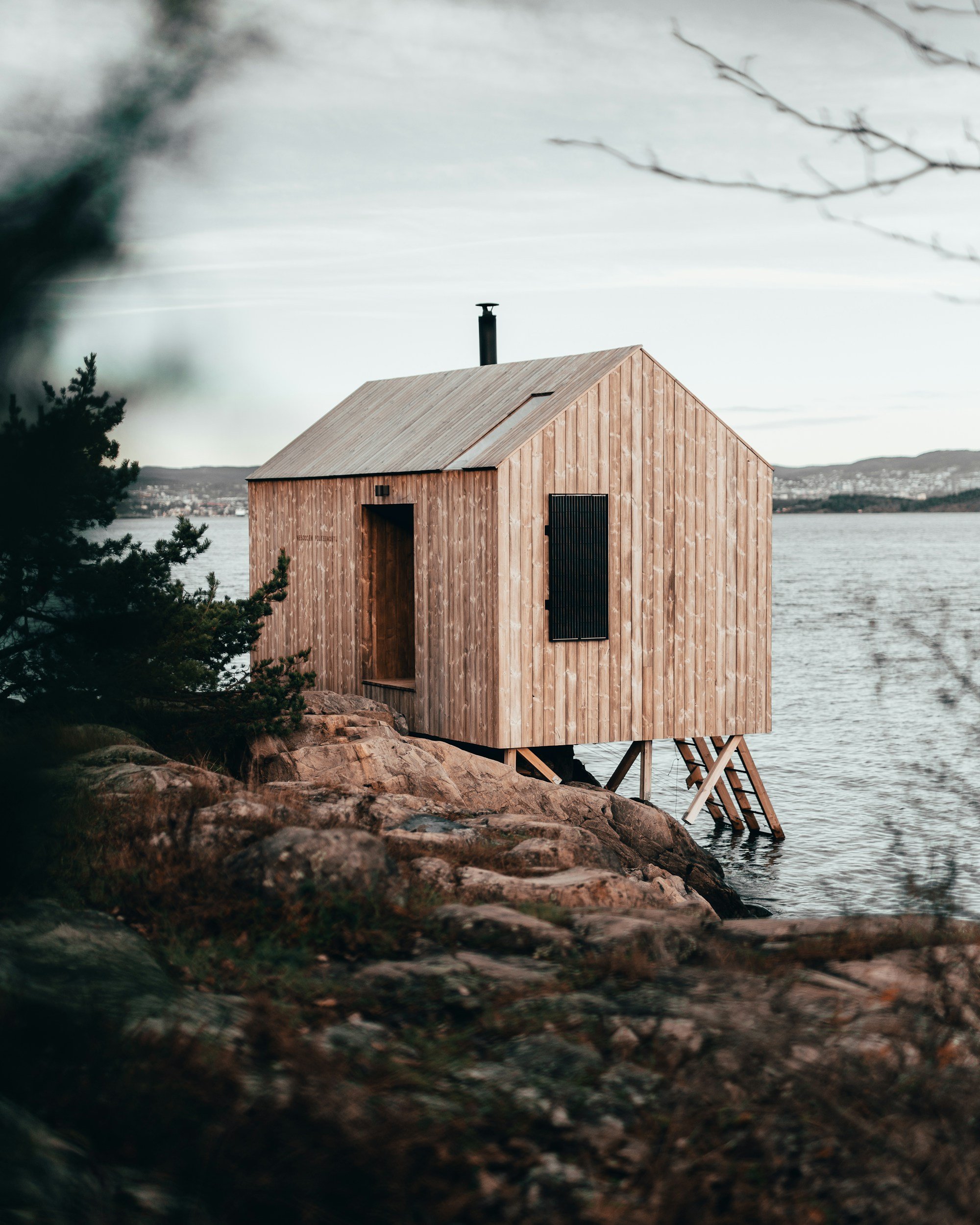 A wooden cabin built on stilts over a body of water, with a ladder leading down to the water, surrounded by natural landscape and trees.