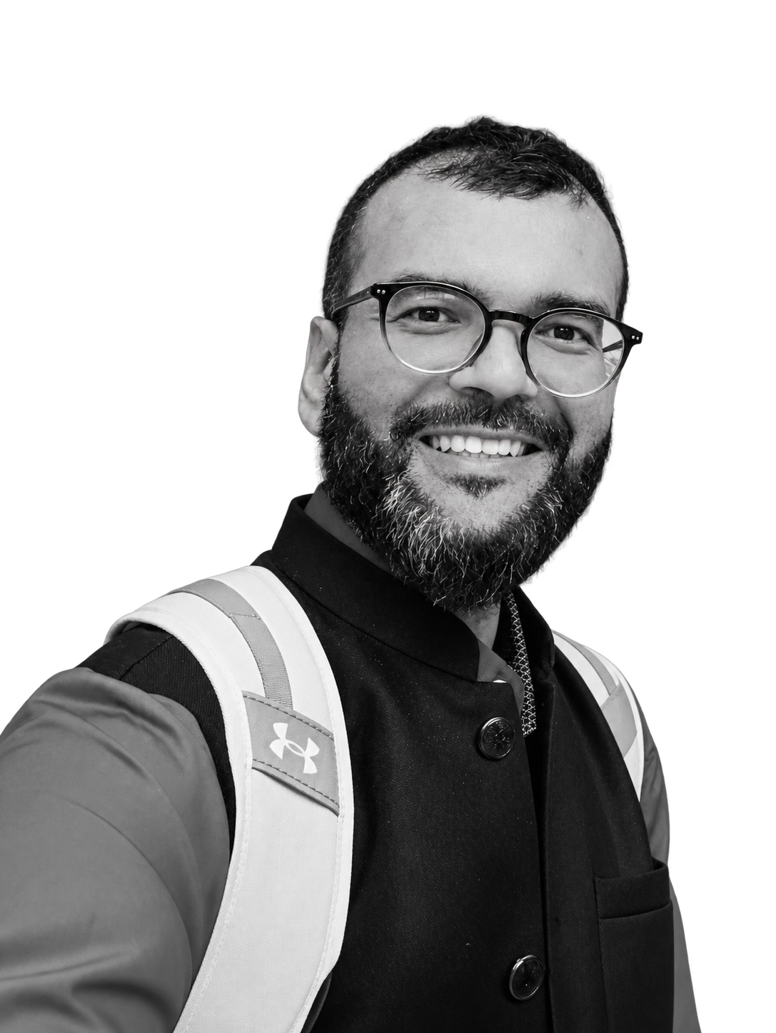 A smiling man with glasses and a beard, wearing a jacket with a backpack, posing for a selfie against a black background.