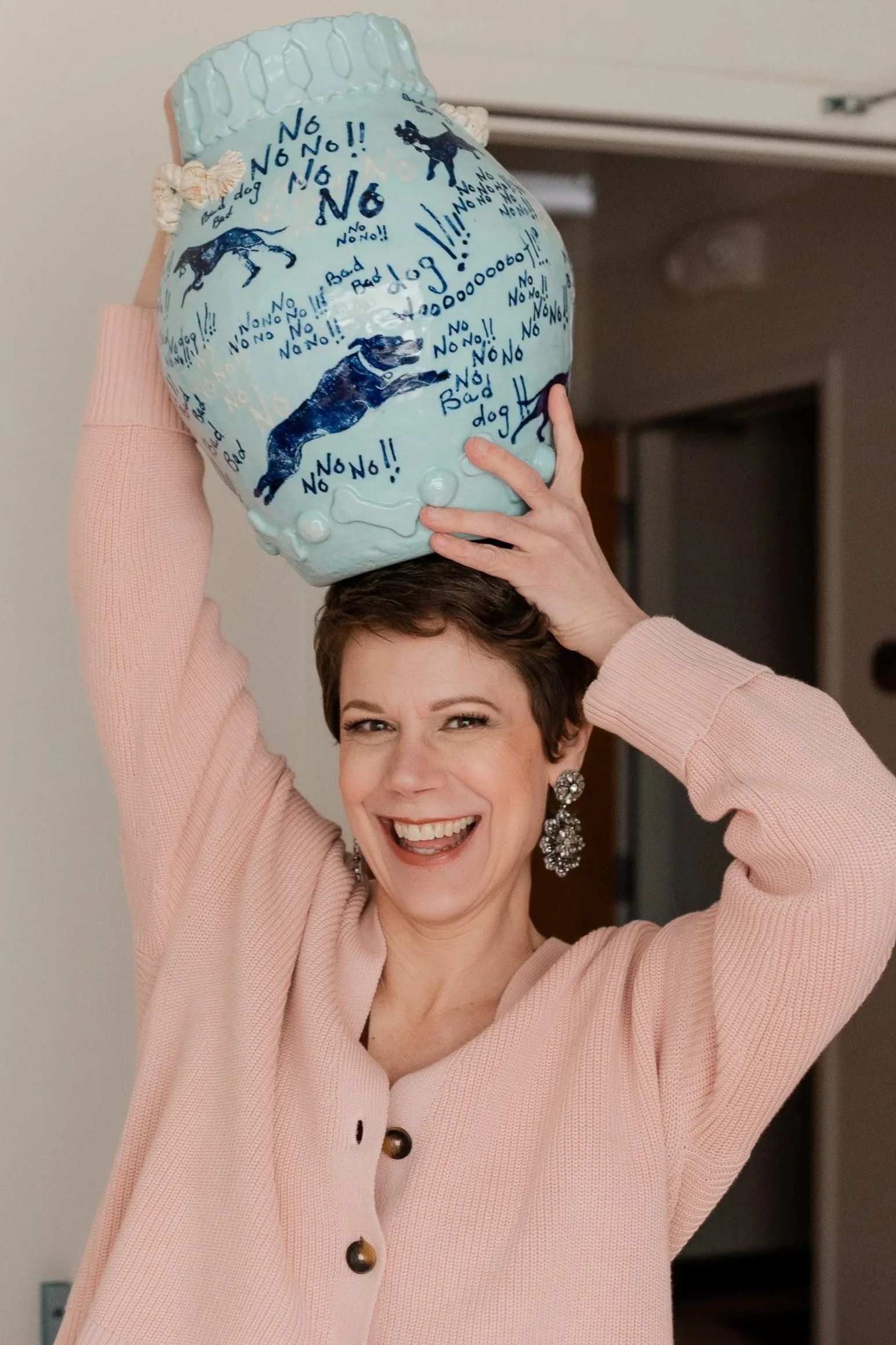 A woman holding a ceramic dog figurine above her head, smiling joyfully. The figurine has various dog-related words and images in blue and black.