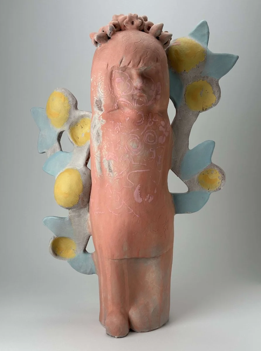 A ceramic sculpture of a human figure with a pink body, standing with a face that has closed eyes. The figure has a crown or head adornment, and large abstract wings with yellow and blue elements extend from its back.