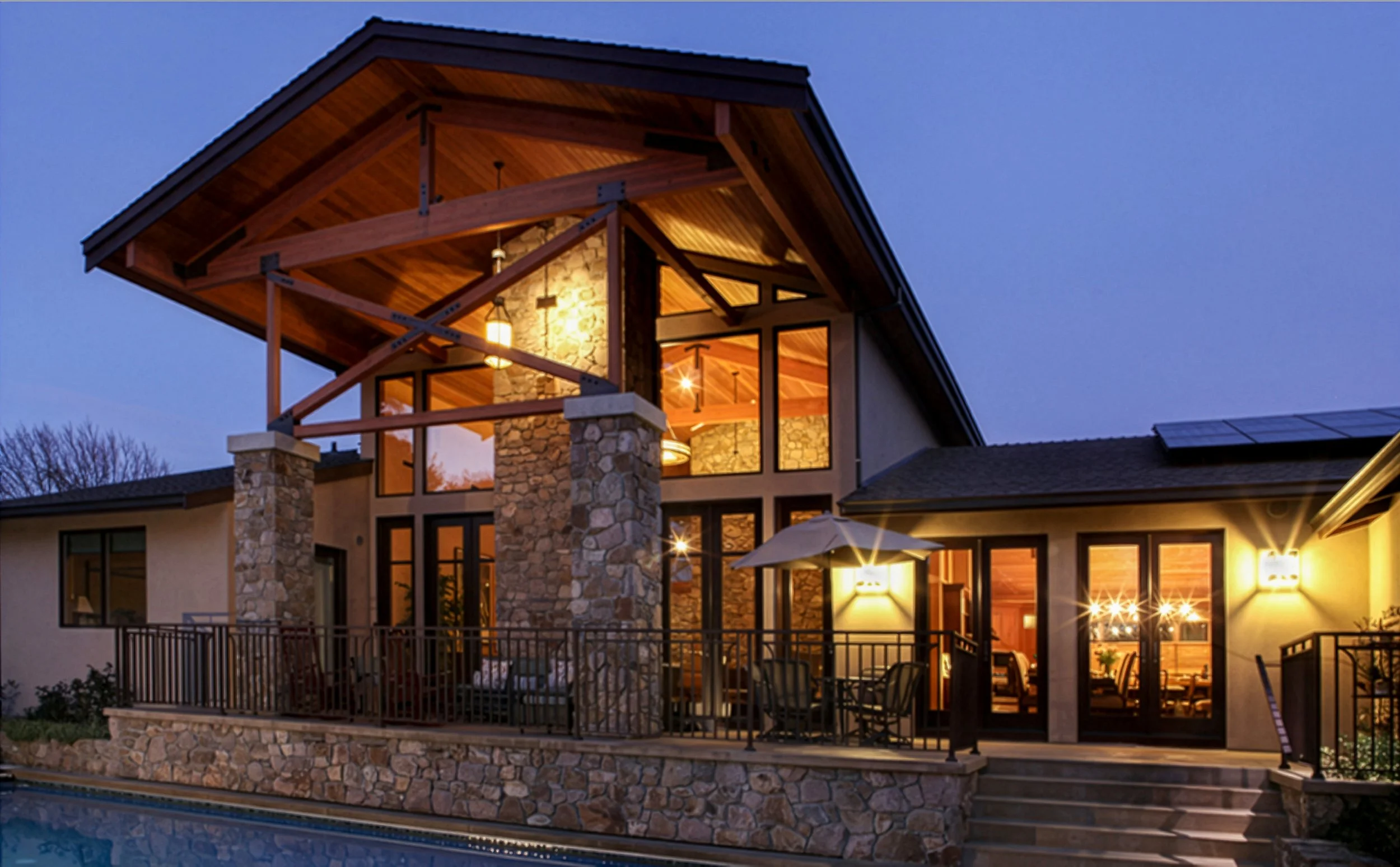 A modern house with large windows, stone and wood exterior, outdoor seating area with an umbrella, and a swimming pool in the foreground, illuminated at dusk.