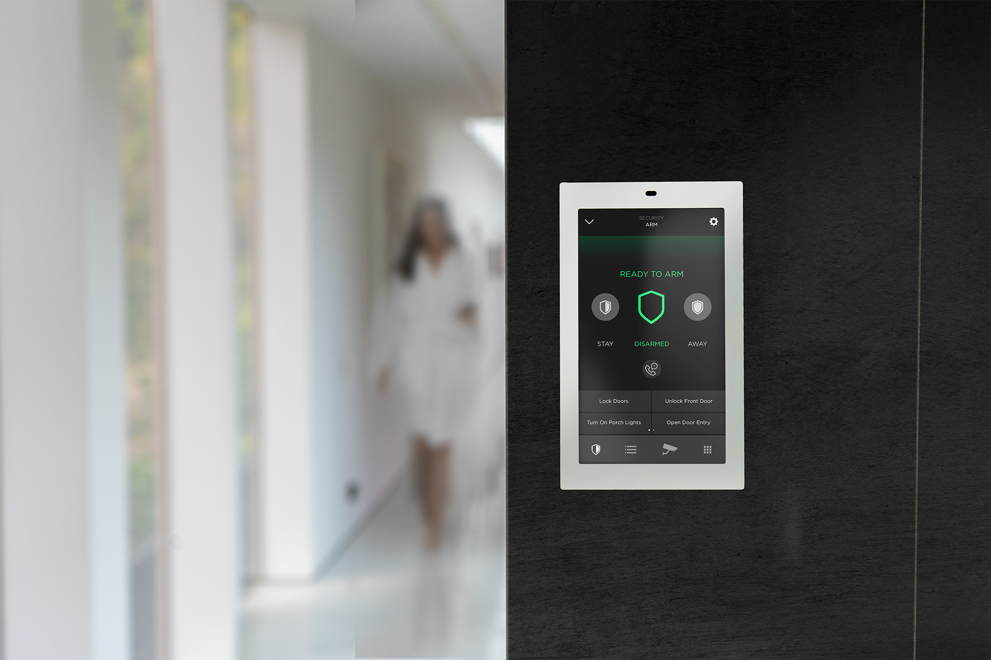 Smart home security control panel mounted on a wall with a blurred woman walking in the background.