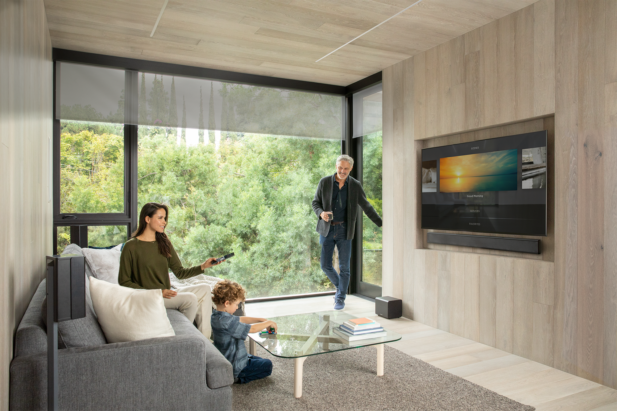A family of three enjoying time in a modern living room with large windows overlooking green trees. A woman is sitting on a gray sofa holding a remote, a young boy is playing with a toy on a glass coffee table, and an older man is entering the room holding a glass, smiling. A large flat-screen TV is mounted on a light wood-paneled wall.
