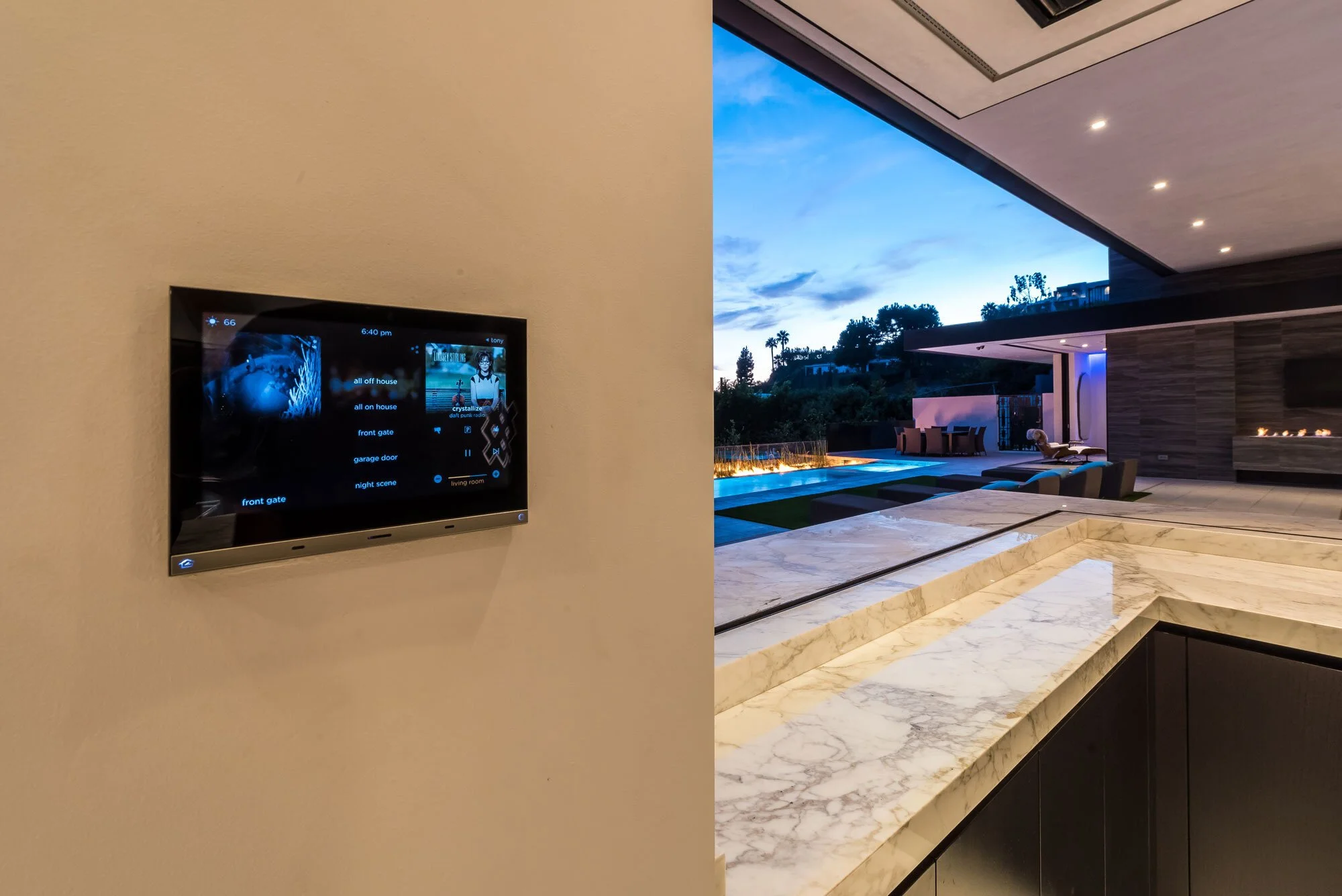 Indoor view of a modern home with a television on the wall showing a remote control app, and a large window revealing an outdoor pool area during dusk with lounge chairs and a fireplace.