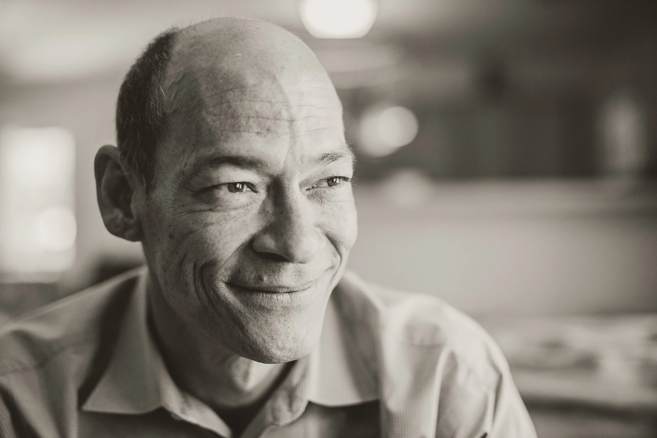 Black and white close-up of a smiling middle-aged man with a receding hairline, wearing a collared shirt, in an indoor setting.
