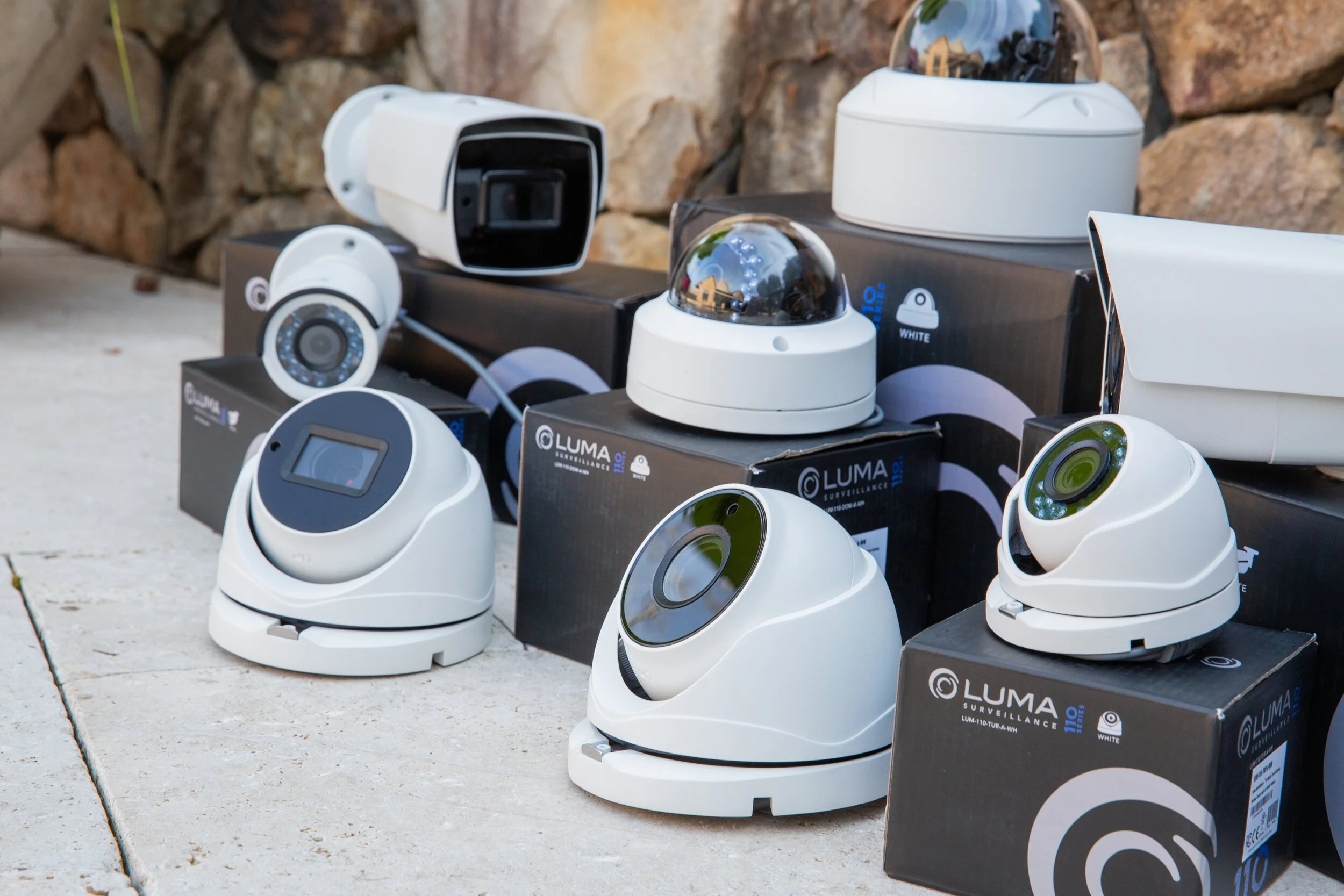 Various models of white security cameras and surveillance devices displayed on the ground and their boxes, with a stone wall in the background.
