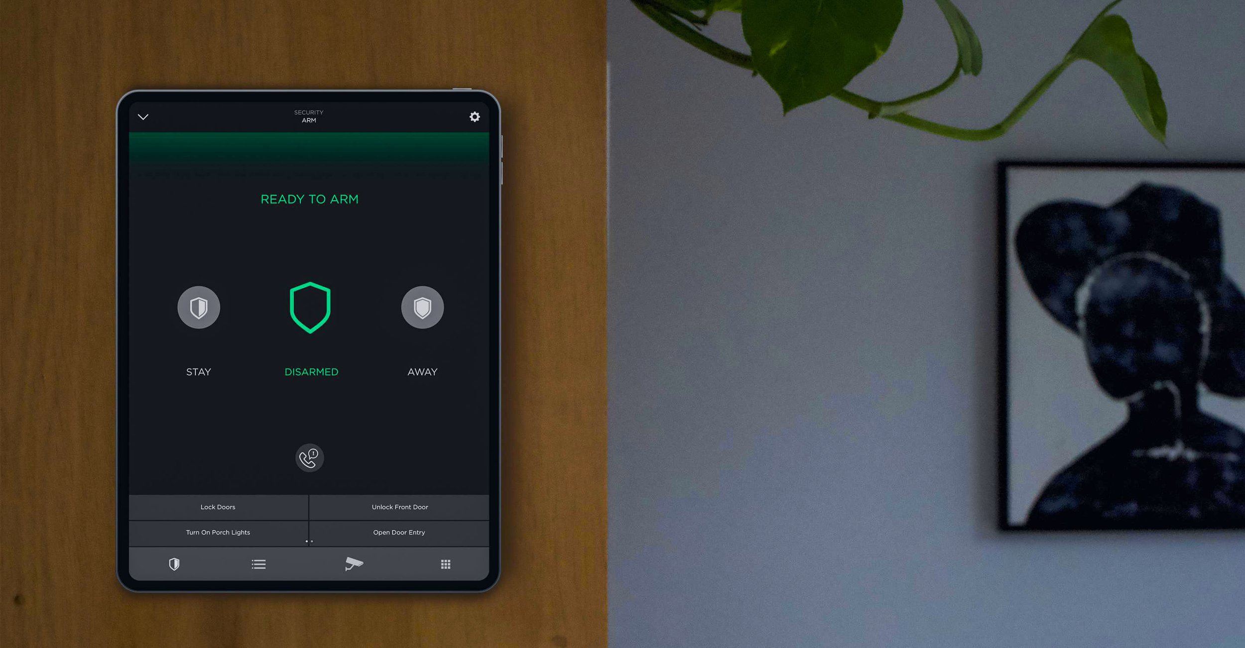 Smartphone displaying home security app with options to stay, disarmed, or away, placed on a wooden surface near a wall with a plant and a framed black and white abstract artwork.