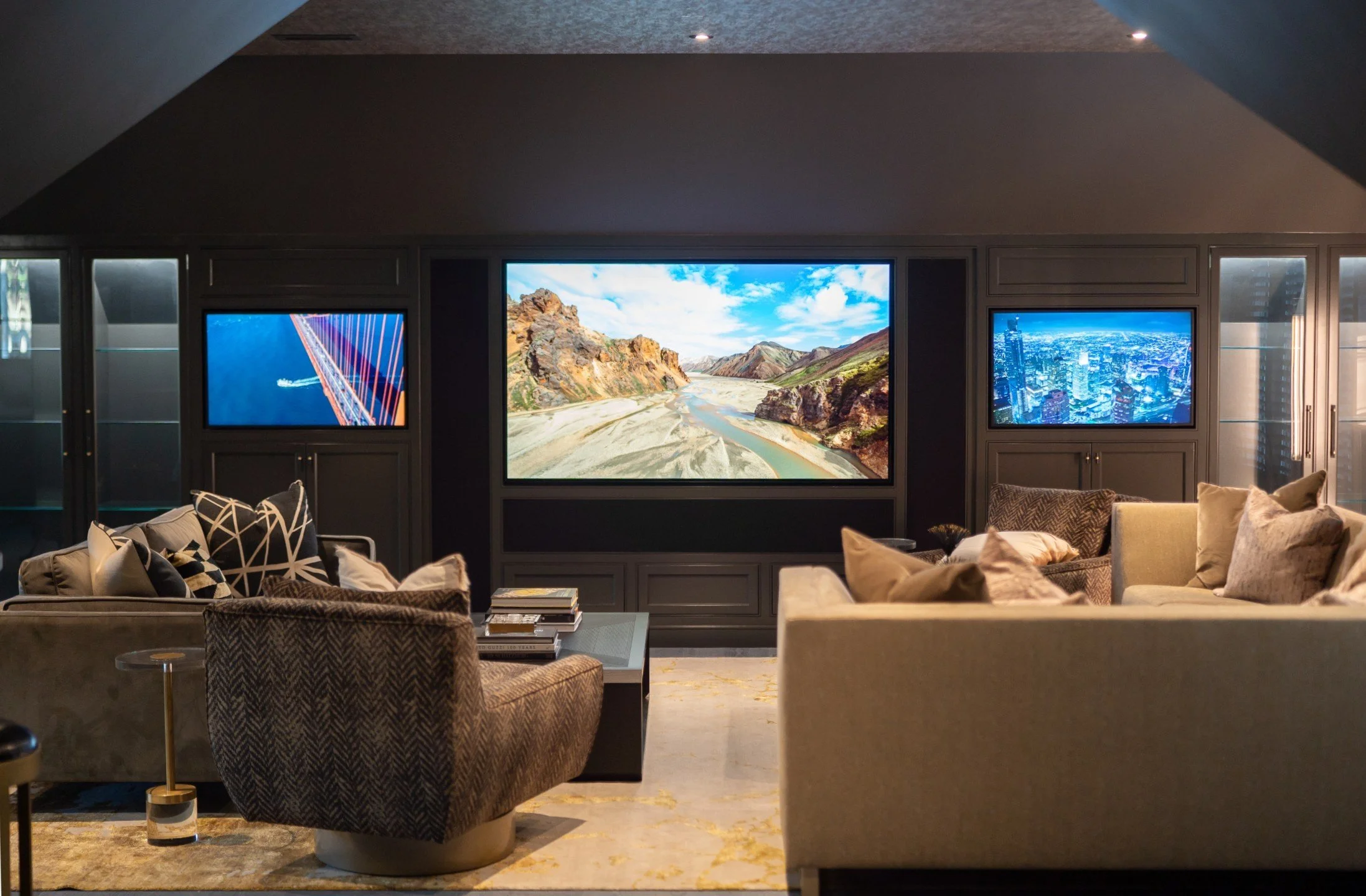 Luxurious living room with three large screens displaying landscapes and cityscape, beige and patterned sofas with pillows, a marble coffee table with books, and modern decor