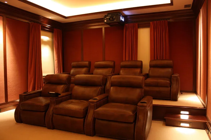 Home theater room with six brown leather recliner chairs arranged in two rows, red curtains, wall-mounted projector, and warm lighting.