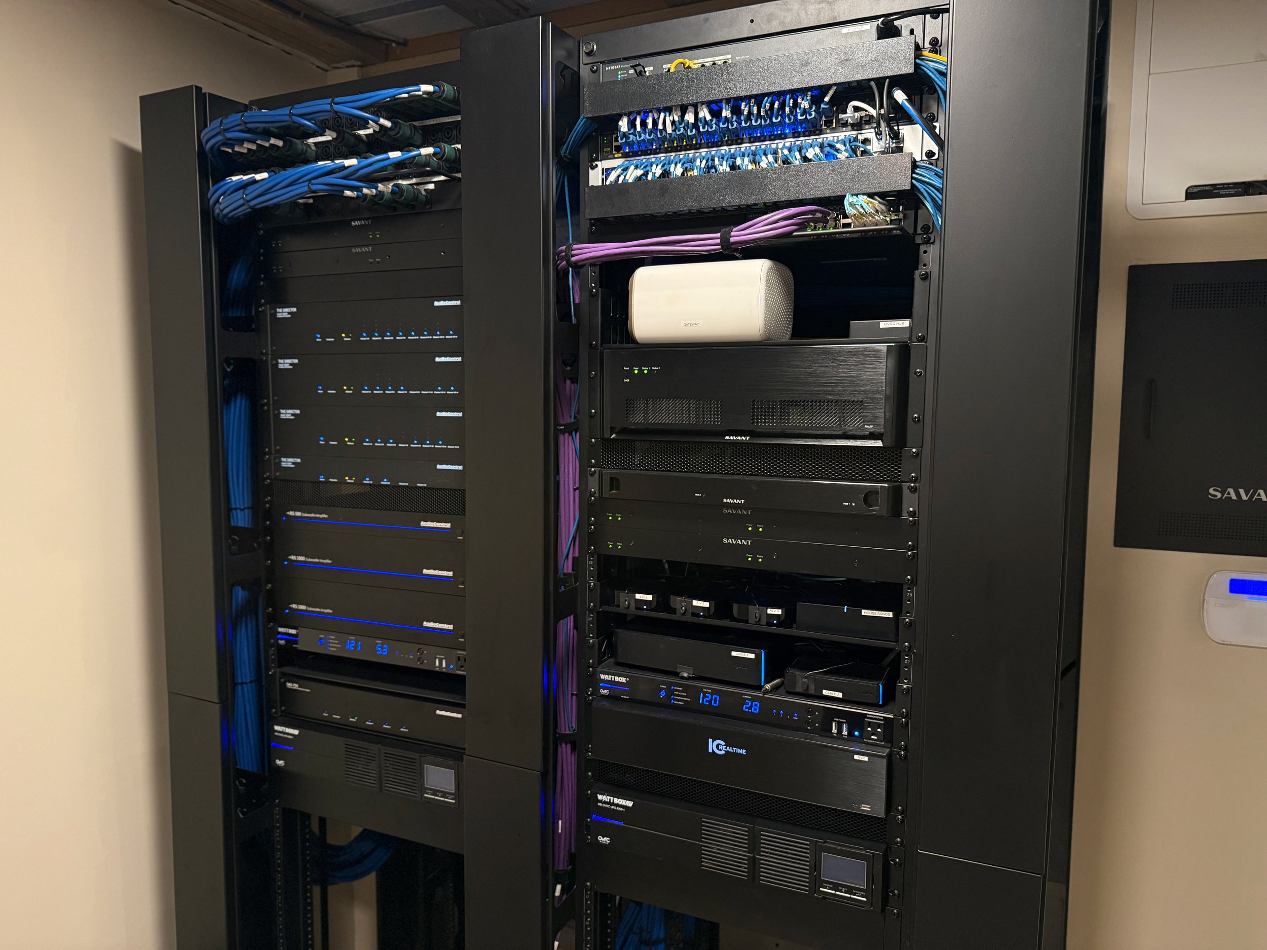 A tall black server rack with open doors, containing various electronic equipment and cables, located in a room with white walls and gray carpet.