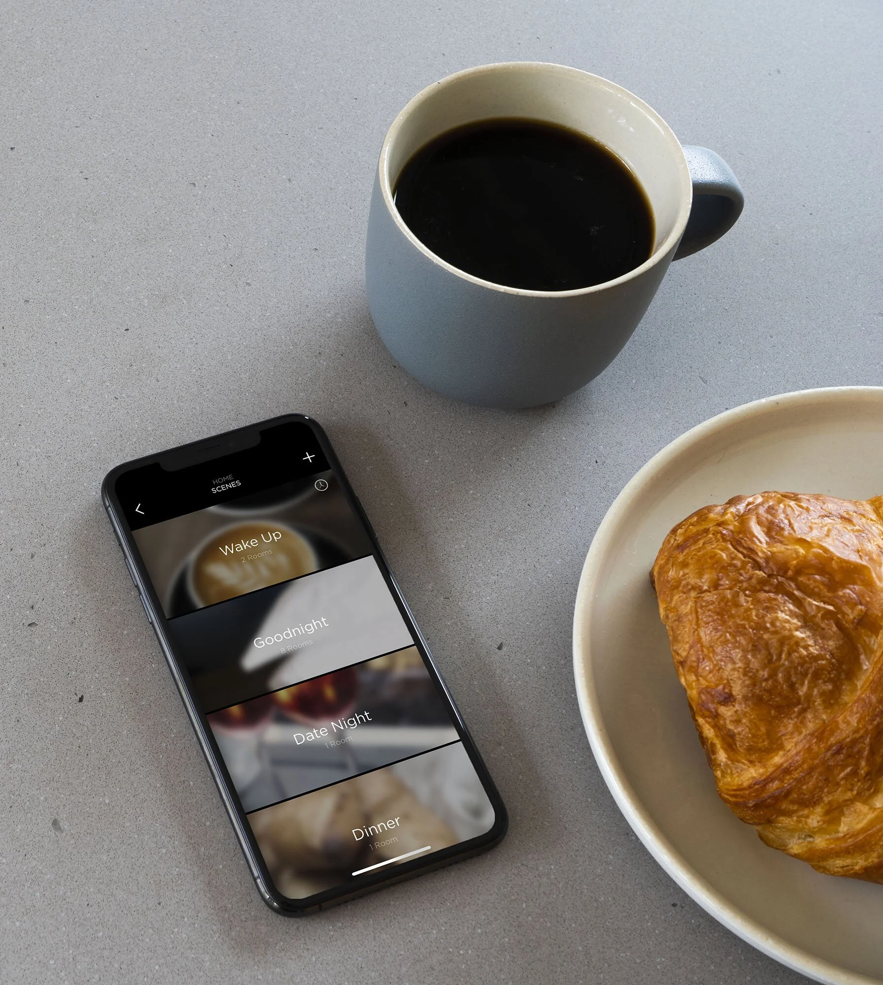 A cup of black coffee, a croissant on a plate, and a smartphone displaying alarm sounds on a gray surface.