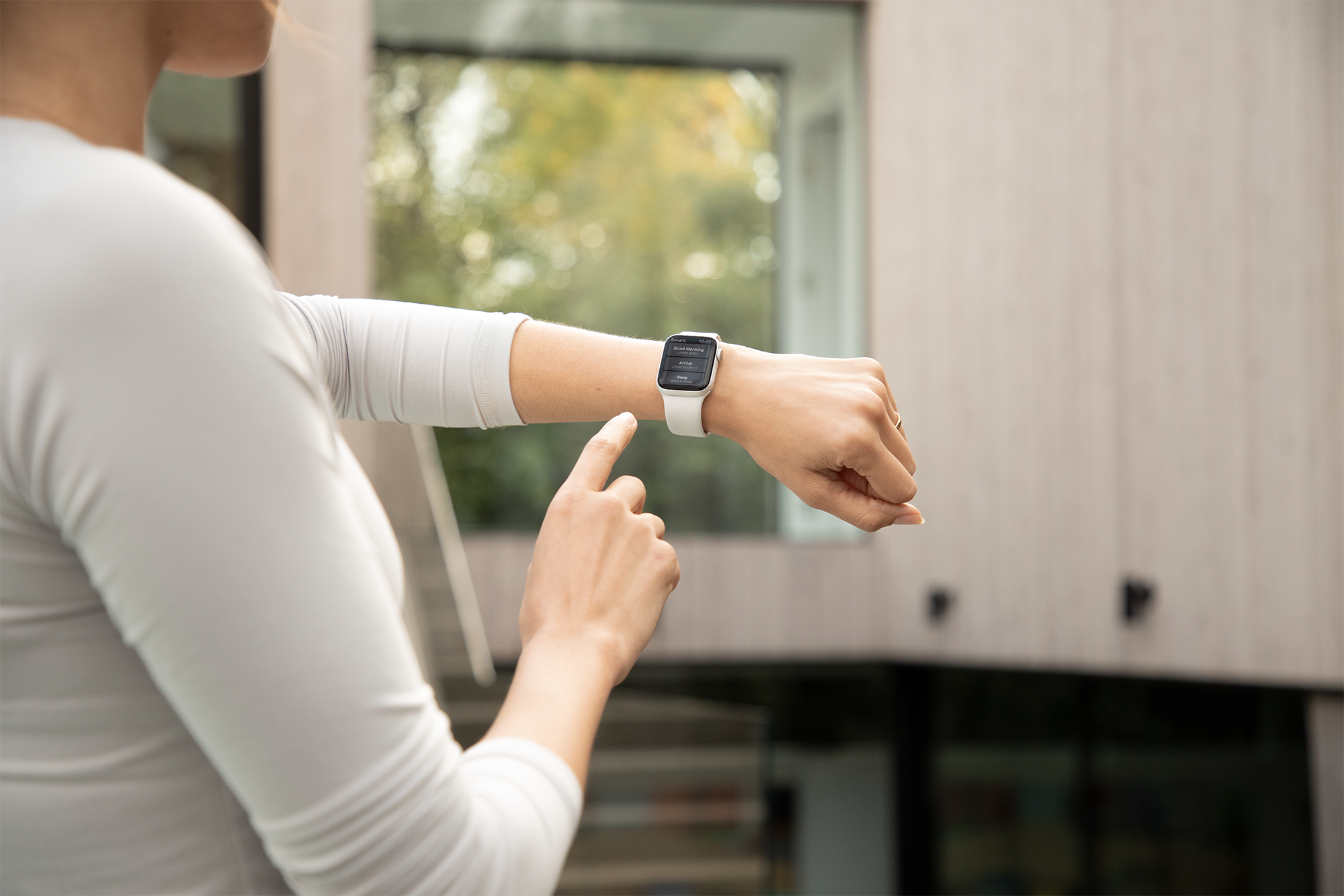 Woman outdoors looking at her smartwatch on her wrist, with her finger pointing at it, in front of a modern house and trees with fall foliage.